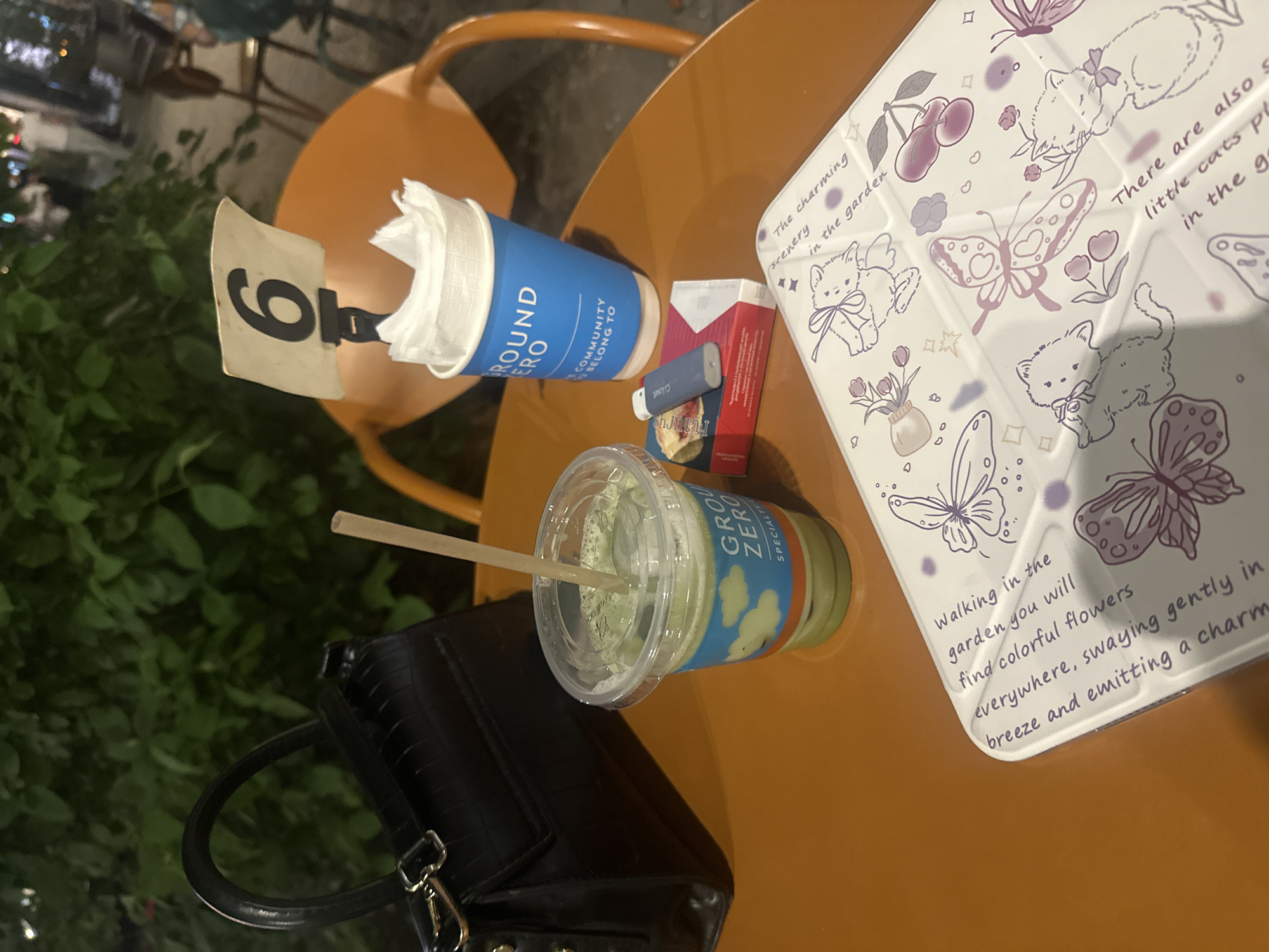 A high-angle shot shows an outdoor table setting. On the table, there is a blue paper cup filled with tissues and another plastic cup filled with a green matcha drink, a red and white cigarette box with a small blue electronic cigarette, and a white tablet cover decorated with cats and butterflies. A black handbag is next to the matcha drink. The number "6" is visible on a white card attached to the back of a chair. The table is orange. The location appears to be an outdoor cafe in Yerevan, Armenia.