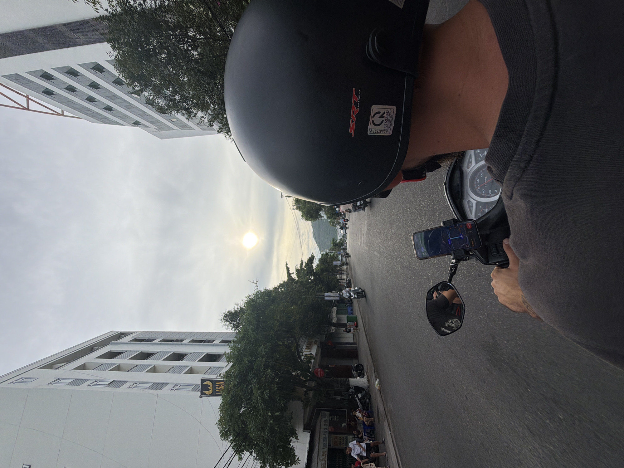 The image shows a first-person perspective of someone riding a scooter in what appears to be a city. The rider is wearing a black helmet with "SRT" and a certification sticker visible. In front of the rider, a smartphone is mounted on the handlebars, displaying a navigation app. The scooter's dashboard is also visible. The road is paved and lined with buildings on both sides. On the left, there is a tall, modern building with balconies and the sign "LASAN" visible. Trees and smaller buildings are also present along the street. Several people are seen sitting and standing on the sidewalk on the left side of the road, and other scooters are in motion on the street. The sky is overcast, with the sun faintly visible, suggesting it might be early morning or late afternoon. The environment suggests a tropical or subtropical urban setting, consistent with coastal Vietnam.