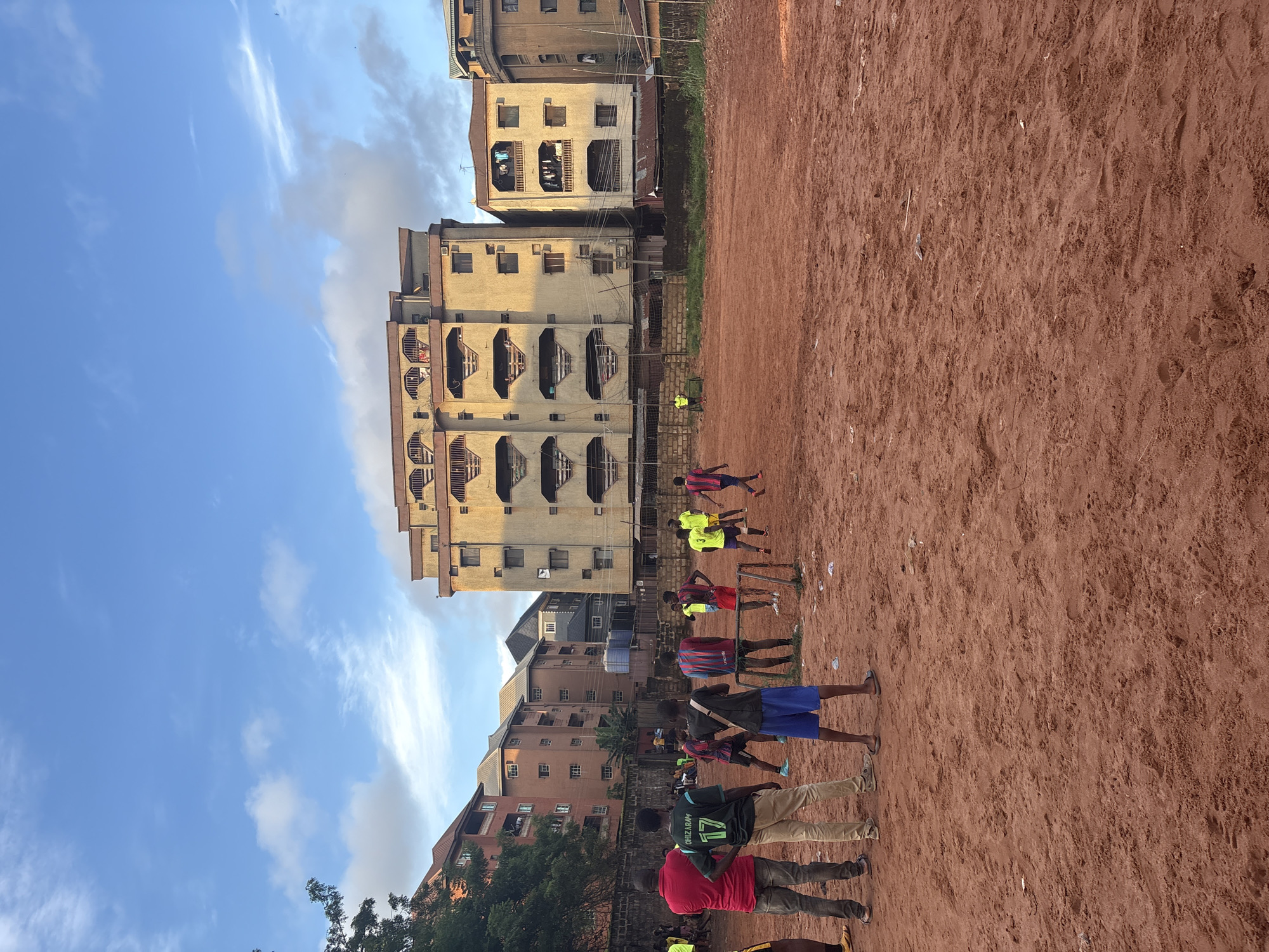 A wide shot captures a group of young men playing soccer on a dirt field in Onitsha, Nigeria. The field is a reddish-brown dirt, marked with a few scattered pieces of litter. A makeshift goal with a net stands on the field, suggesting an informal game. In the background, multi-story apartment buildings rise, some with balconies displaying laundry. The sky above is a bright blue, dotted with white clouds. The people are dressed in casual clothing, some wearing soccer jerseys. The overall scene depicts a common urban recreational activity in a densely populated area.