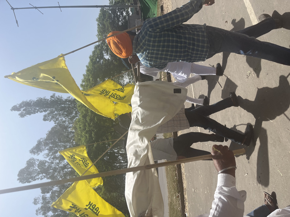 A group of men are walking down a paved road in Sahibzada Ajit Singh Nagar, India, carrying a white cloth banner and waving yellow flags with Punjabi writing on them. The men are wearing traditional Indian clothing, including turbans and long shirts. The road is lined with trees and there are several people in the background. The men are walking in a single file line, with the man at the front carrying the banner. The man at the back of the line is waving a yellow flag. The sky is blue and the weather is sunny. The man at the back of the line is wearing a blue plaid shirt and blue jeans. He is carrying a bamboo pole and a yellow flag. The banner is being held by the man in front of the group, who is wearing a white shirt and a blue turban. The man behind him is wearing a white shirt and black pants. The man at the front of the line is wearing a white shirt and blue jeans. The man in the middle of the line is wearing a white shirt and white pants. The man in the back of the line is wearing a blue plaid shirt and blue jeans. The banner is a white cloth sheet that is being held up by the men. The yellow flags are being waved in the air. The group is walking down a paved road in Sahibzada Ajit Singh Nagar, India.