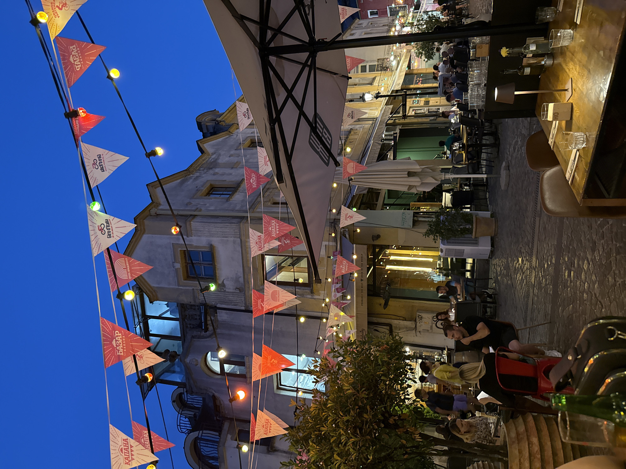 This image captures a lively evening scene at an outdoor restaurant or cafe, likely in Plovdiv, Bulgaria, given the text visible on the decorative flags. The sky is a deep twilight blue, indicating it is either dusk or early evening. Strings of colorful lights and triangular flags are draped across the scene, creating a festive atmosphere. The flags feature Cyrillic text, including "КАПАНА" (Kapana, a well-known creative district in Plovdiv) and "БАЗАР" (Bazaar), along with what appears to be "Giro d'Italia" branding on some.
People are seated at tables, engaged in conversation and enjoying their evening. Some are dressed casually, while others appear to be dining or socializing. The overall mood seems relaxed and convivial. In the foreground, part of a table setting with glasses and bottles is visible, suggesting a close-up perspective from someone seated at the establishment. The architecture in the background suggests an urban setting with traditional European influences, characterized by stone buildings and balconies. The presence of an umbrella and potted plants further enhances the outdoor dining ambiance. The lighting from the string lights and from within the buildings creates a warm, inviting glow, contrasting with the darkening sky.