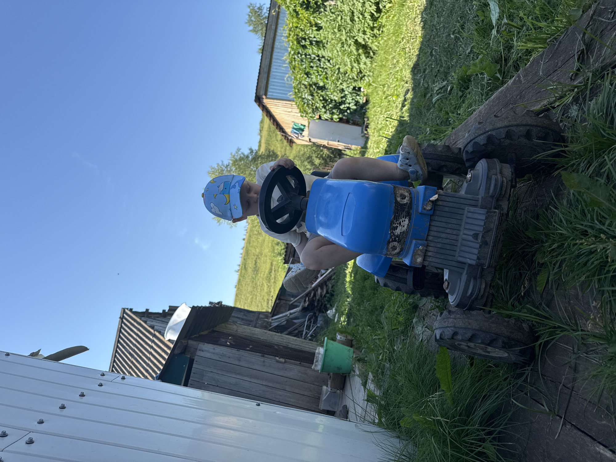 A young boy wearing a blue cap sits on a blue toy tractor in a rural outdoor setting. The boy is holding the steering wheel. The tractor is on a wooden surface with grass and weeds around it. In the background, there is a wooden building with a corrugated metal roof, and a dense green hedge. Beyond the hedge, a grassy hillside is visible under a clear blue sky. The scene appears to be in a village or countryside location, possibly in Ляльшур, Russia.