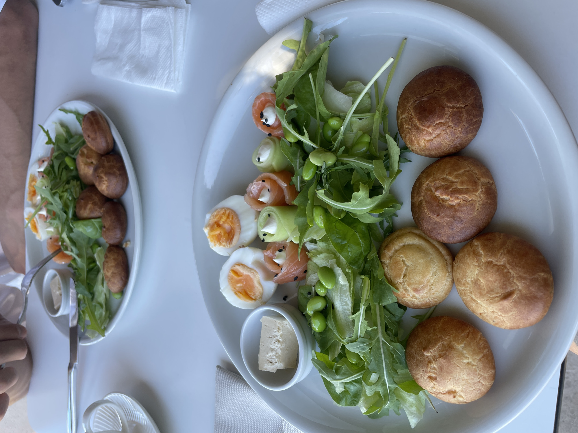 Two plates of food are on a white table. The plate in the foreground contains a salad with rocket, edamame, and smoked salmon rolls stuffed with cream cheese and cucumber. There are also two halves of a hard-boiled egg, a small bowl with a cube of cheese, and four small, round, golden-brown rolls. A person's hand is visible in the upper left corner, holding a fork and knife over the other plate. This second plate, slightly out of focus and in the background, appears to hold a similar salad with rocket and orange segments, as well as several more of the small, round rolls. White napkins are stacked to the right of the foreground plate. The scene suggests a meal, likely breakfast or brunch, being served at a restaurant or cafe in Rafailovići, Montenegro, given the context provided.