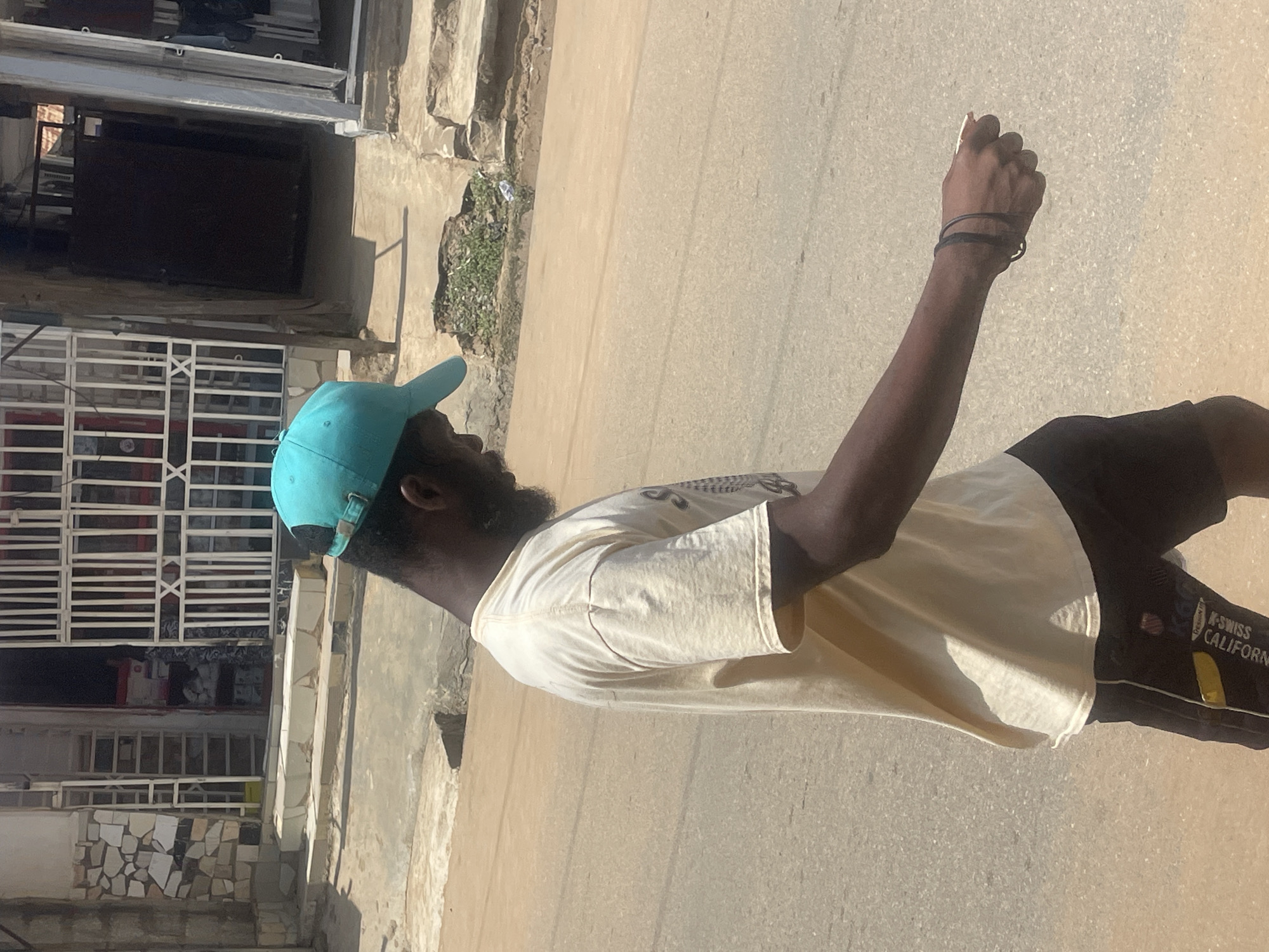 A man with dark skin, wearing a light-colored t-shirt and dark shorts, stands on a paved surface, possibly a street. He has a turquoise baseball cap on and appears to be holding a cigarette in his right hand, which is raised. His left arm is by his side. He has a beard and is seen from the side, looking to his left. The background shows a building with barred windows and a rough stone facade. There is also a patch of green vegetation growing from a crack in a stone wall. The ground is a light beige asphalt. The lighting suggests it is daytime, with a strong shadow cast by an unseen object to the left. On the side of his shorts, there is text that appears to read "K-SWISS CALIFORNIA". The scene seems to be in an urban environment, with architectural elements typical of Abuja, Nigeria. The man's posture and the cigarette suggest a moment of pause or a casual activity.
