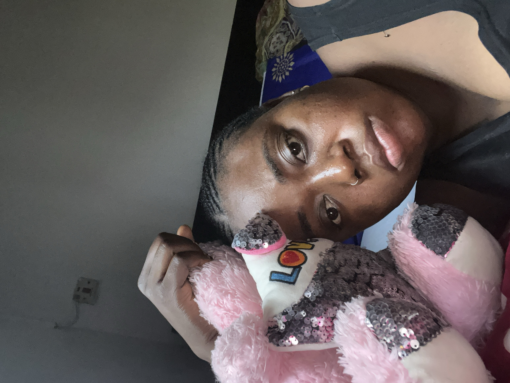 The image captures a person, likely a young adult female, lying down and looking directly at the camera. She has dark skin, braided hair styled close to her head, a nose piercing, and an ear piercing. She is wearing a dark-colored t-shirt.
In her embrace, she holds a plush toy that is predominantly pink and adorned with silver sequins. The word "LOV" is partially visible on the toy's front, suggesting the full word is "LOVE."
The setting appears to be indoors, with a plain, light gray wall serving as the primary background. A fragment of patterned blue fabric with a golden, sun-like emblem is visible behind her head. A white wall socket with visible wiring is on the gray wall to the left. The lighting is soft and ambient, with no harsh shadows, indicating an indoor environment, possibly during the day or with artificial light. The person's expression is neutral and direct, suggesting a calm or contemplative mood. The photo appears to be a selfie taken in a domestic setting, consistent with the location context of Abuja, Nigeria.