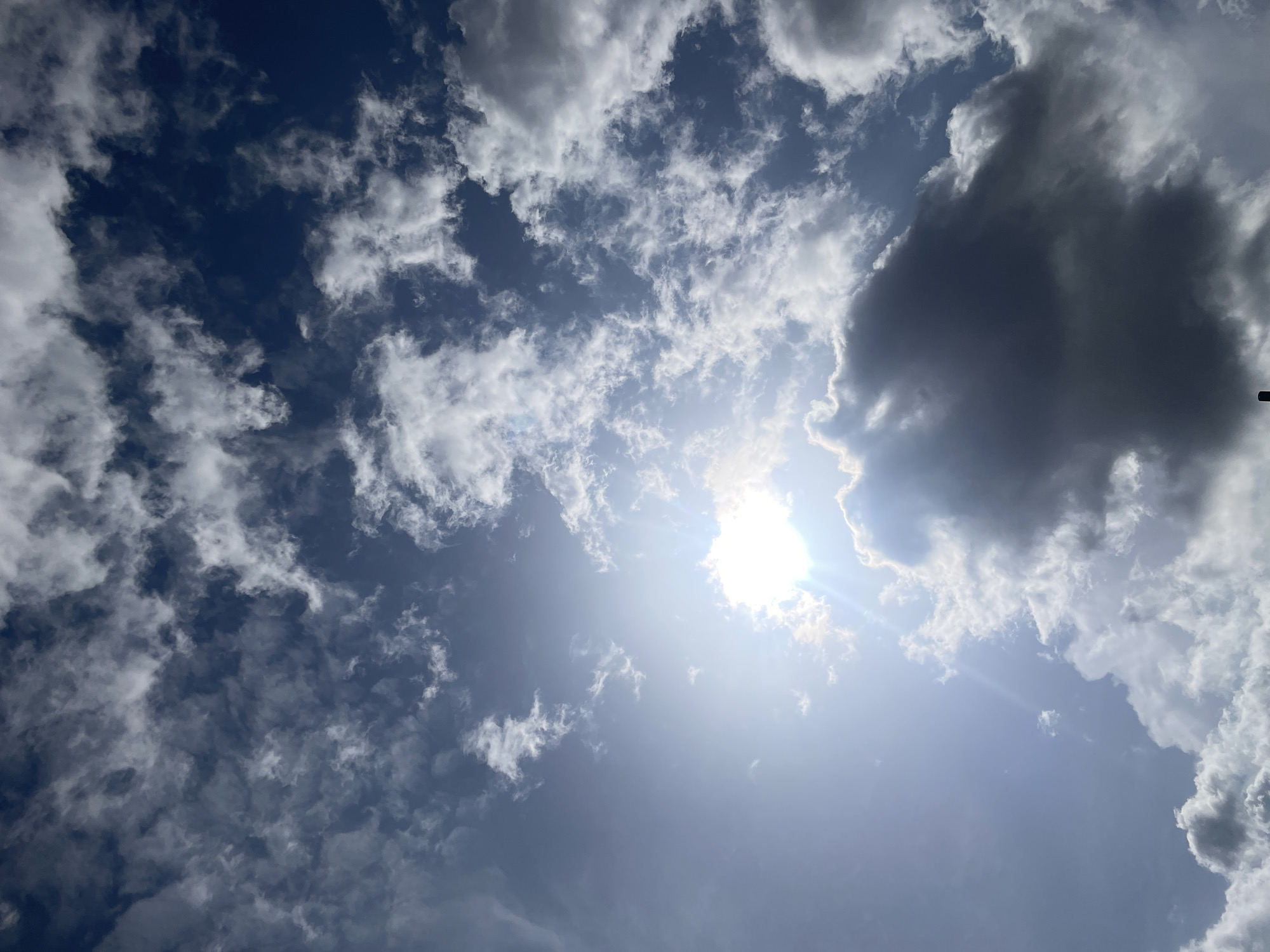The image shows a daytime sky in Jos, Nigeria, with a bright sun partially obscured by white clouds and a large, dark gray cloud. The sky is a deep blue color. The sun is located in the lower center of the frame and emits bright rays. The clouds vary in size and shape, with some appearing thin and wispy, while others are dense and puffy.