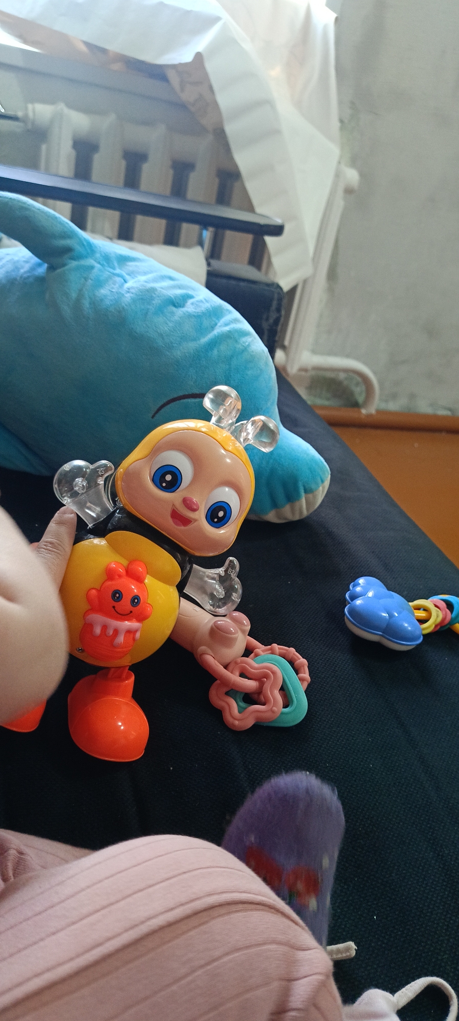 An overhead shot shows a close-up of a baby's toy bee with a yellow body and orange feet. The bee is next to a large blue plush dolphin. The toy bee has large blue eyes, clear plastic antennae, and a red, cupcake-shaped decoration on its body. A baby's hand is visible, reaching towards the toy. To the right of the bee, a blue cloud-shaped rattle is visible, along with a pink and teal teething toy. In the lower right corner, a baby's foot wearing a purple sock with a red strawberry pattern is visible. The background shows a dark, textured surface, possibly a couch or carpet, with a hint of a white cloth and a radiator in the upper portion of the image. The scene appears to be indoors. There is no information in the image to suggest the location is the city of Хохлы, Russia.
