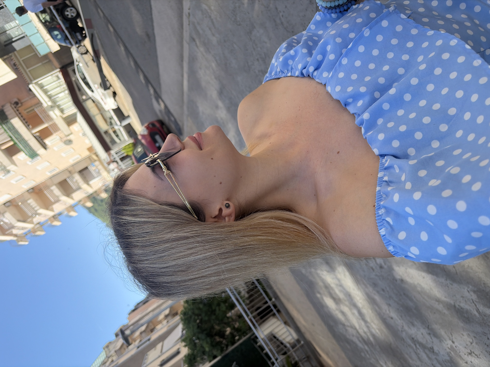 A sideways outdoor portrait features a young woman seen in profile, looking upwards towards a bright blue sky. Her blonde to light brown hair is visible, framing her face. She wears gold-rimmed sunglasses perched on her head or side of her face. A small dark stud earring is visible in her left ear. She is dressed in a light blue, off-the-shoulder top adorned with white polka dots. A blue beaded bracelet is visible on her right wrist in the upper right corner of the frame.
The background depicts an urban environment, likely in Budva, Montenegro, under clear and sunny daytime conditions. To the left, several multi-story buildings with light-colored facades are visible, along with parts of a street where cars are parked and driving. Some green foliage from trees can also be seen. The woman appears calm, with a serene expression, possibly posing or simply enjoying the sunny weather. The immediate foreground shows a grey, textured surface, which could be pavement or a balcony floor. There is no visible text in the image.