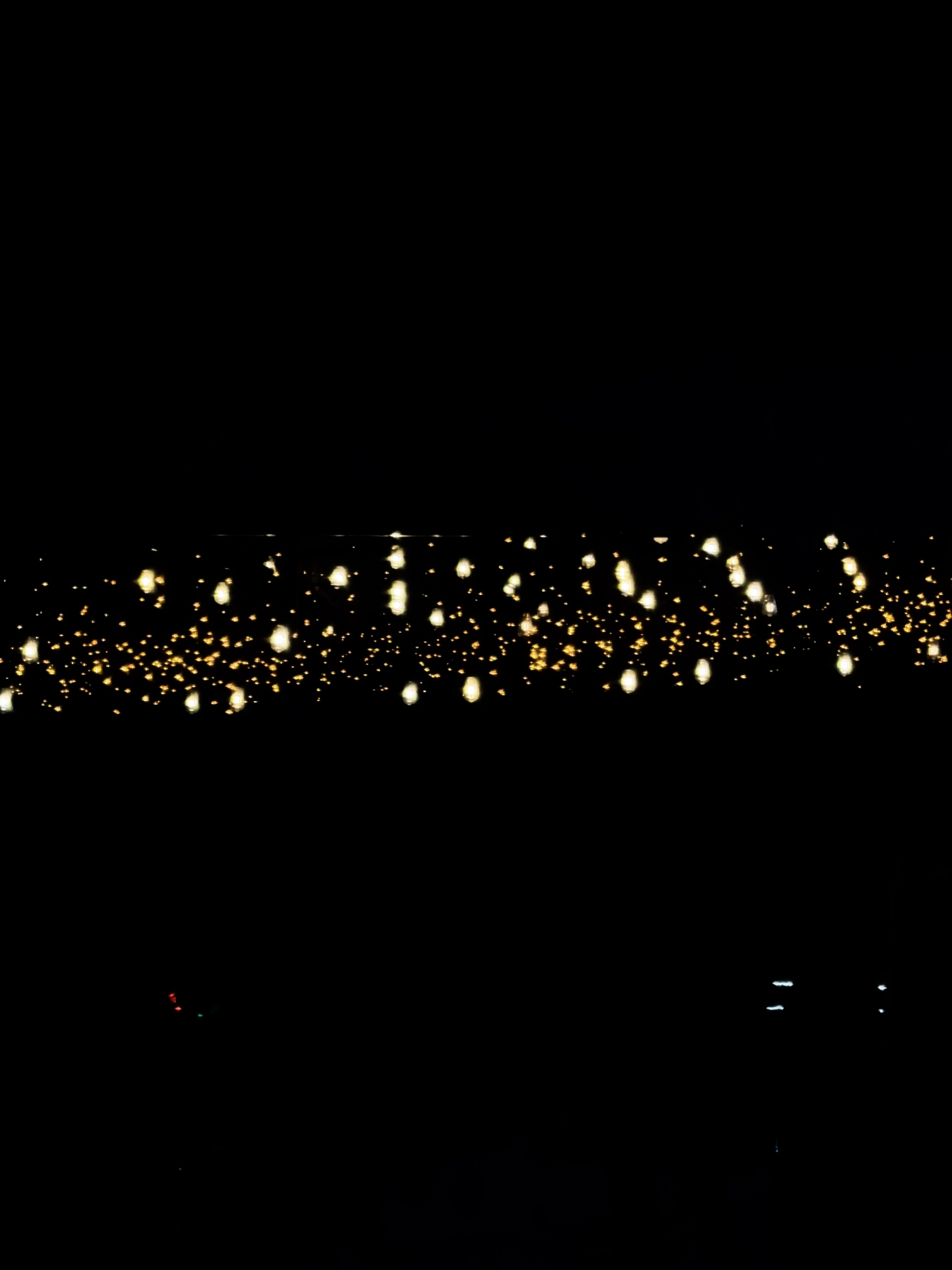 The image shows a dark background with a horizontal band of blurred golden bokeh lights. The bokeh lights are small, circular, and glow brightly, creating a festive or magical effect. The rest of the image is entirely black, with a few faint, vertical white lines or streaks at the bottom right. There are no discernible people, objects, or specific environmental features visible in the image. The image does not provide any information about its location, including Fergana City, Uzbekistan.