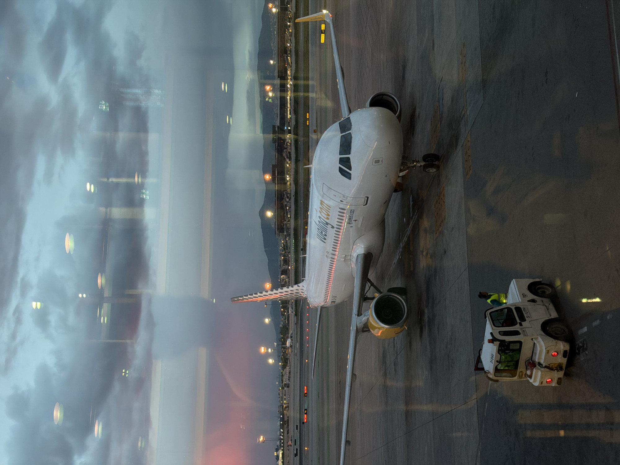 The image shows an airplane parked at Barcelona–El Prat Airport, which is located near Sant Boi de Llobregat, Spain. The Vueling airplane is being serviced by a tow truck. The photograph was taken from inside the airport through a window, as evidenced by the reflection of the interior and the exterior scenery. The time of day is likely dusk or dawn, as the sky is reflecting in the window with a range of colors from light to dark. Beyond the plane, the cityscape stretches into the distance, with hills visible in the background. Runway lights and airport infrastructure are also apparent.
