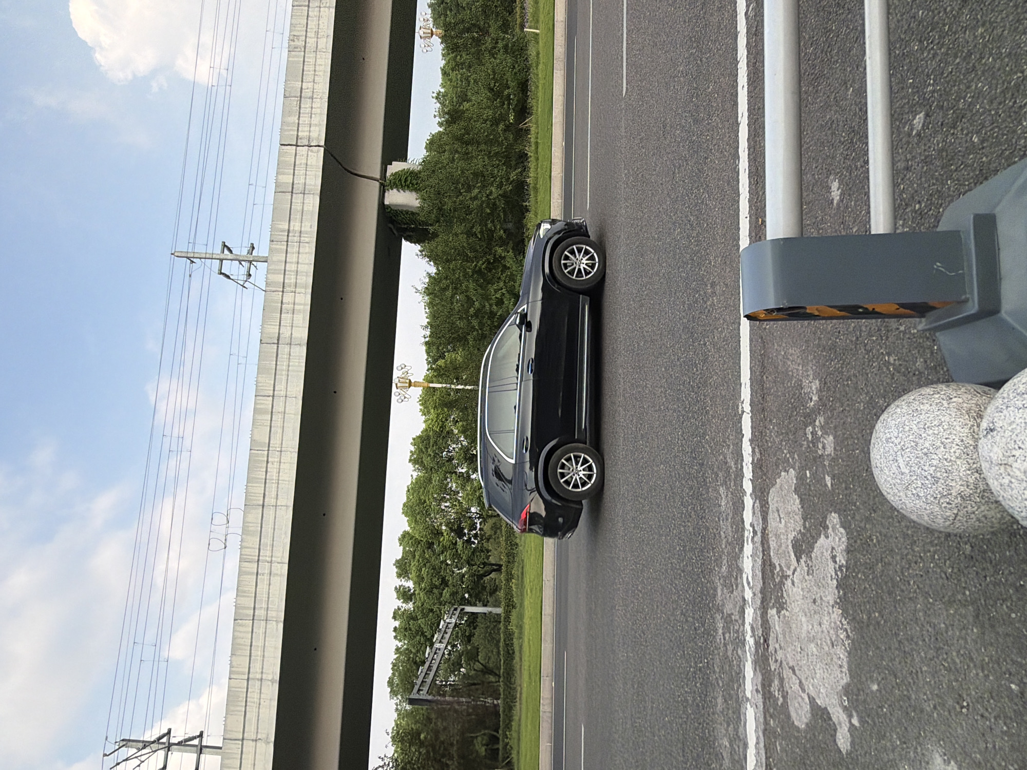 A black Mercedes-Benz GLE SUV is captured in profile driving on a paved road under an elevated concrete overpass. The overpass supports overhead electrical wires and appears to be part of a highway or major roadway. Dense green trees line the side of the road beyond the overpass, with a clear sky and scattered clouds above. In the foreground, parts of a gray guardrail with yellow and black reflective strips and two speckled stone spheres are visible. The scene is likely set in Jiangjin District, Chongqing, China, given the characteristic infrastructure and landscape.