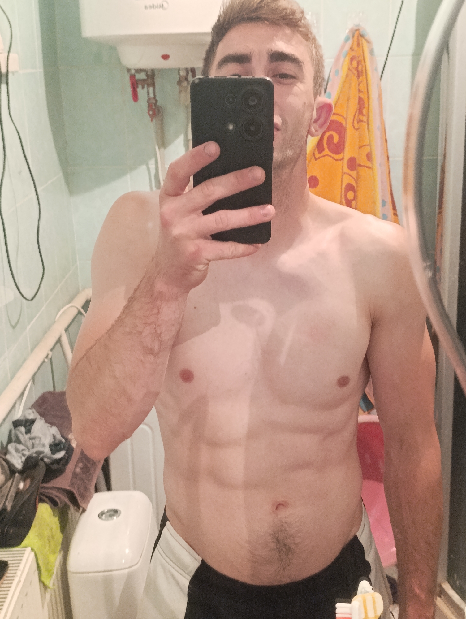 The image shows a shirtless man taking a mirror selfie in a bathroom. The man has blond hair and is holding a black smartphone in his right hand. He is wearing black and white shorts. The bathroom appears to be small and tiled with light green tiles. A water heater is visible on the wall behind him, as well as a towel rack with an orange towel. Other objects in the bathroom include a toilet, some laundry, and a radiator. The lighting is dim and the image has a green tint. The location is not specified in the image, and therefore cannot be determined.