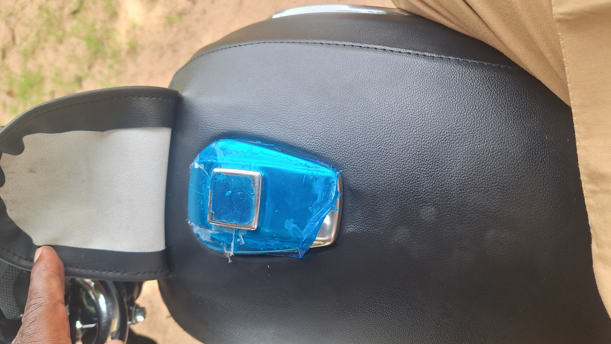 The image shows a close-up of a black motorcycle seat. A flap on the left side of the seat is open, revealing a white fabric lining. In the center of the seat, a motorcycle key is covered in blue plastic wrap. The key has a silver metallic square on it. The seat is textured, resembling leather. A person's hand is visible on the left side of the image, pointing towards the open flap. Part of a tan colored garment is visible on the right side, draped over the seat. The background appears to be outdoors, with blurred dirt and green vegetation, suggesting a rural or semi-rural setting. The lighting indicates it might be daytime. The overall impression is of a motorcycle being prepared or maintained. There is no visible text in the image.