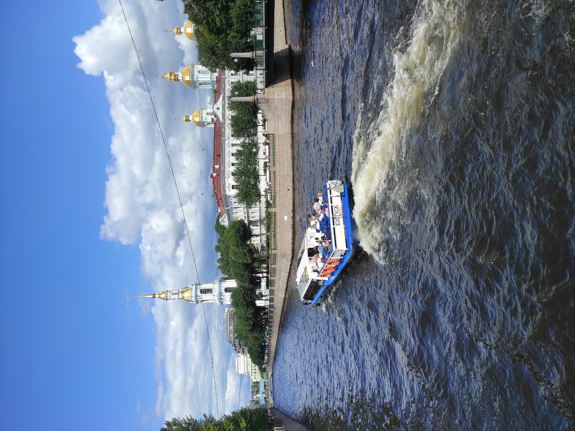 This is a medium-shot, eye-level, outdoor, vertical photograph of a boat tour on a canal in Saint Petersburg, Russia. The boat, which is white with a blue bottom, is moving away from the viewer, leaving a white wake in the dark blue water. There are many people on board the boat, some sitting and some standing. The boat has a sign that says "КНИГА" which translates to "book" in Russian. In the background, there are several buildings with gold domes, a white bell tower with a gold spire, and trees lining the canal. There are also clouds in the blue sky. The architecture suggests this is likely the area around the St. Nicholas Naval Cathedral, a prominent landmark in Saint Petersburg.