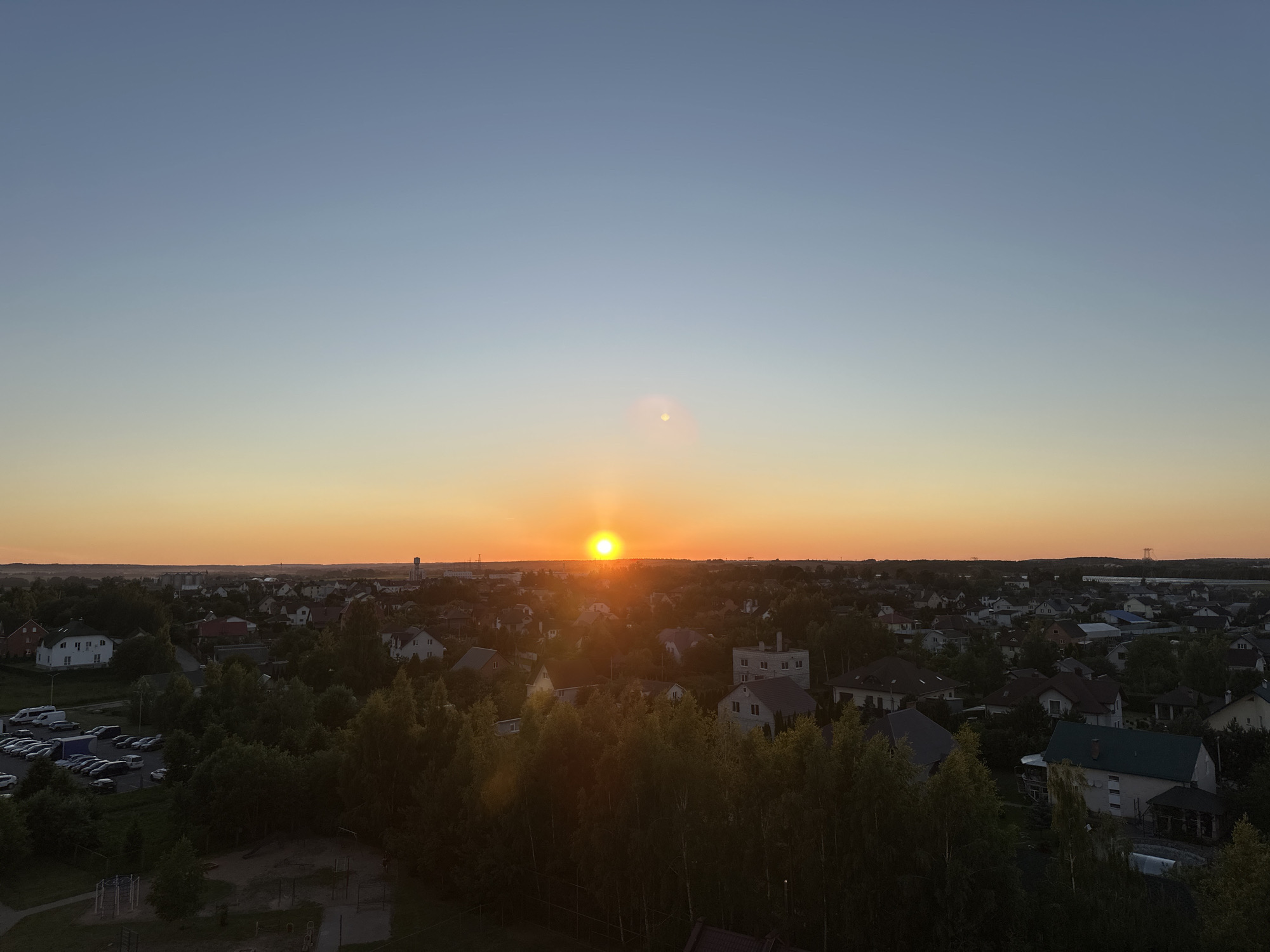 The image captures an aerial view of a residential area in the Shchomyslitski Sel'ski Sovet, Belarus, during sunset. The sun is low on the horizon, casting a warm, orange glow across the sky and illuminating the landscape. The scene is dominated by a dense collection of houses with various roof colors, interspersed with lush green trees. In the foreground, a dark area suggests a park or playground with visible structures. To the left, a parking lot is filled with numerous cars. The horizon is dotted with distant buildings and a tall tower, hinting at a more urbanized area beyond the immediate residential zone. The overall atmosphere is tranquil, with the soft light of dusk settling over the community.