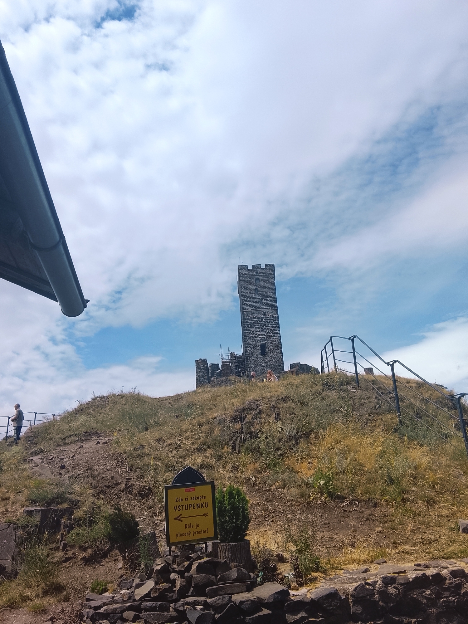 A tall, dark stone tower stands on a grassy hill under a cloudy sky. A metal railing lines a path leading up the hill towards the tower. A yellow sign with black text and an arrow points to the right, indicating a ticket purchase and paid area. In the foreground, rocks and greenery cover the ground. A person stands near the railing on the left side of the frame, looking out over the landscape.