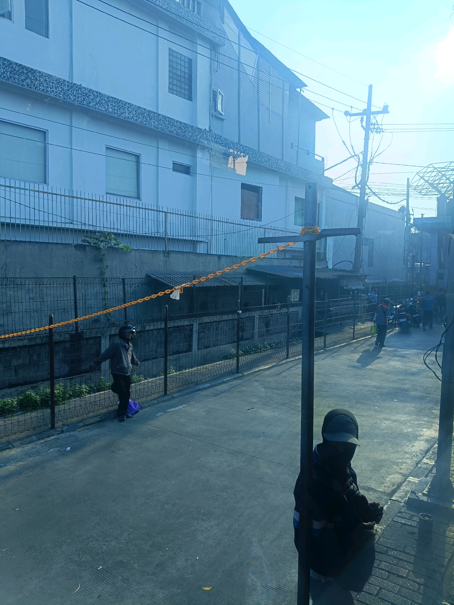 The image depicts a street scene in Kedungrejo, Indonesia, bathed in bright sunlight. A person wearing a helmet and a gray jacket is walking on the left side of the street, pulling a purple bag. In the foreground, another person is partially visible, wearing a dark hooded jacket and a cap, facing away from the camera. The street is paved and lined with fences. On the right side of the street, several motorcycles are parked, and other individuals are present. Behind the street, there is a multi-story building with windows and a fence. Power lines and utility poles are visible overhead. The scene appears to be an outdoor urban environment.