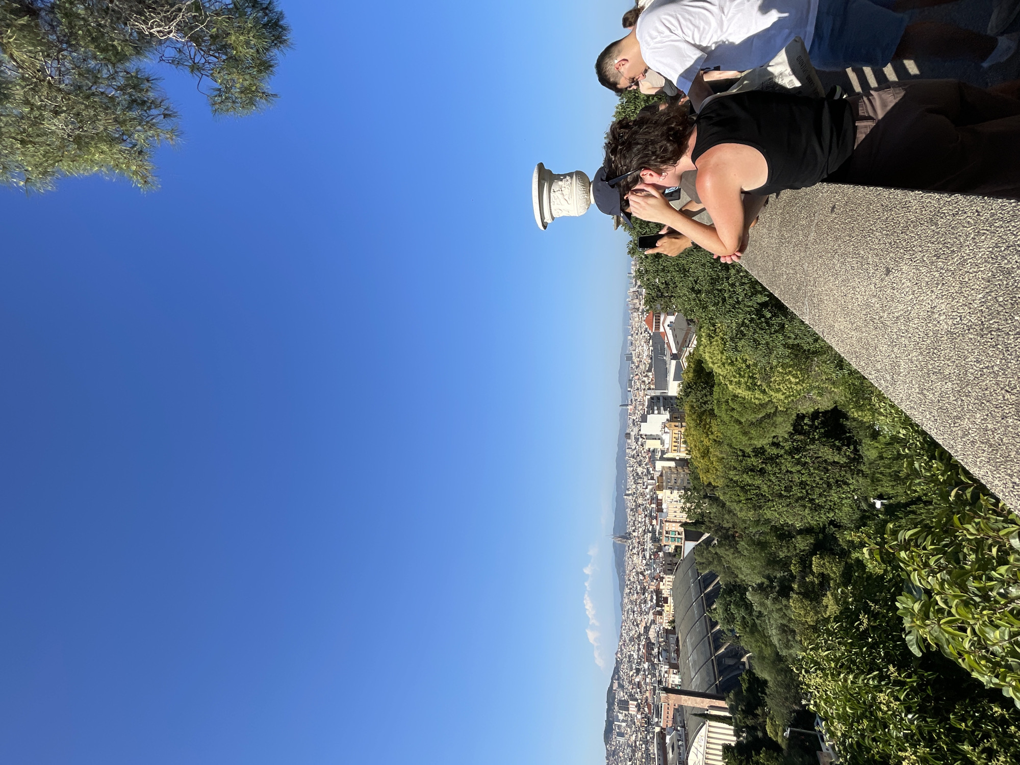 In Barcelona, Spain, from a high vantage point, a panoramic view of the city unfolds under a clear blue sky. In the foreground, a textured stone wall runs diagonally across the frame. Several people are gathered along the wall, observing the cityscape. A woman with dark, curly hair, wearing a black tank top and brown shorts, leans on the wall, holding up a smartphone to take a picture. A man in a white t-shirt and blue shorts stands to her right, also looking out at the view. Further in the distance, the dense urban landscape of Barcelona stretches out, with numerous buildings of varying architectural styles and heights. Among the buildings, a prominent Gothic or Neo-Gothic style cathedral spire, likely the Sagrada Familia, is visible on the horizon, silhouetted against a hazy mountain range. Lush green trees and vegetation are interspersed throughout the city and in the foreground, adding to the scenic beauty of the view. The overall impression is one of a tourist enjoying a picturesque vista of Barcelona on a sunny day.