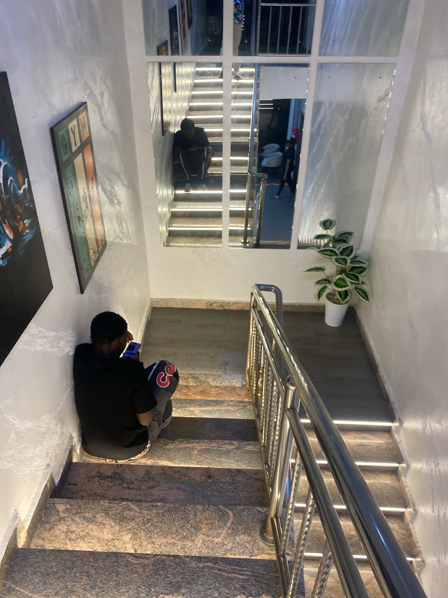 In Barden Yana, Nigeria, a young man wearing a black t-shirt and patterned trousers sits on a granite staircase, looking down at his mobile phone. He is positioned on the lower steps, leaning against a white wall adorned with textured paint and two framed pieces of art. A metal handrail runs alongside the stairs, which are illuminated by embedded lights. Across the staircase, a mirror reflects another individual on a higher flight of stairs, also engaged with a phone. A potted plant with green and white leaves sits near the top of the stairs.