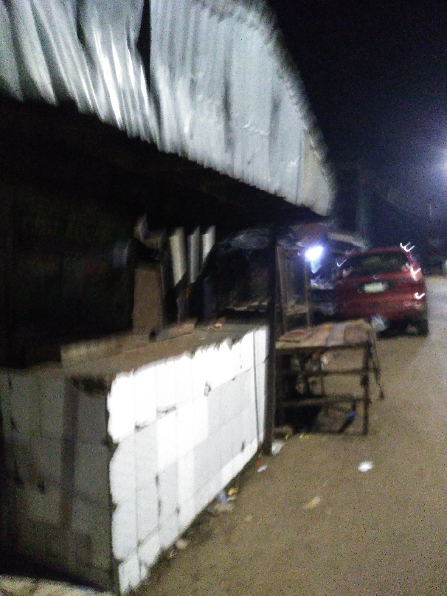 The image depicts a dimly lit street scene at night in Jos, Nigeria. In the foreground, a white tiled structure, possibly a counter or a market stall, is visible. Behind it, a dark, covered structure with a corrugated iron roof extends into the background. To the right of the stall, a metal rack or shelving unit is present. Further in the background, a red car is parked on the street, with its taillights illuminated. The street appears to be paved and somewhat worn. Debris is scattered on the ground in front of the stall and around the car. The overall impression is that of a roadside market or street vending area in a somewhat neglected urban environment.