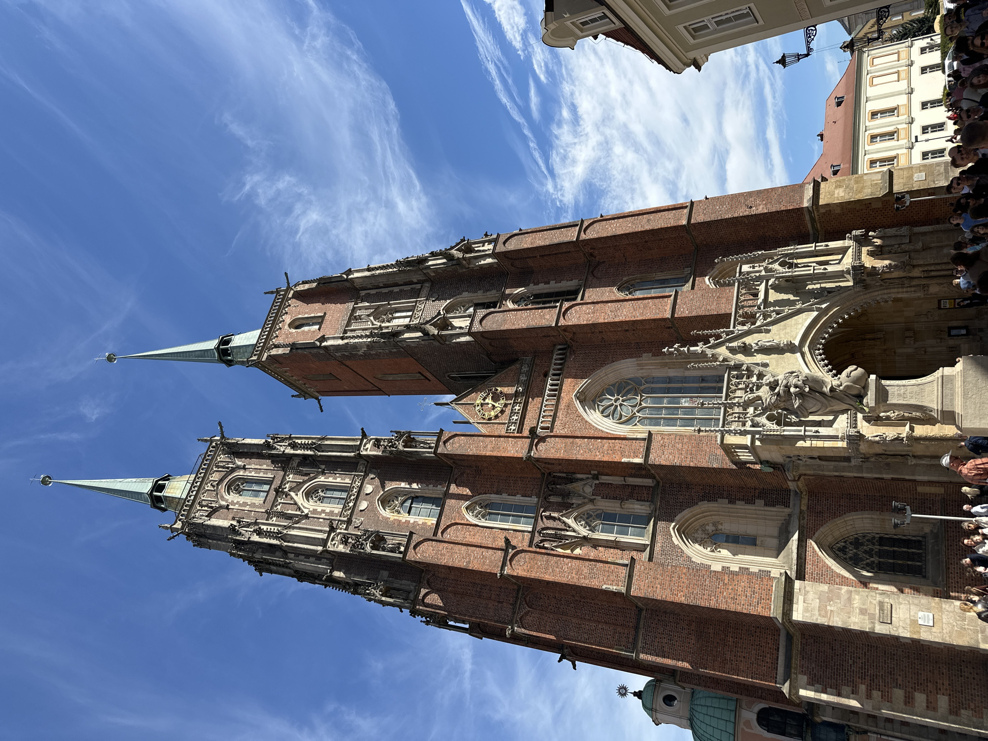 This is a low-angle shot of St. John the Baptist Cathedral in Wrocław, Poland. The cathedral is a large, red-brick Gothic building with two prominent spires that reach towards the blue sky with wispy clouds. Architectural details, including windows, arches, and sculptures, are visible on the facade. A crowd of people is visible near the cathedral, suggesting a tourist attraction. Other buildings are visible, indicating the cathedral is located in a dense urban area.