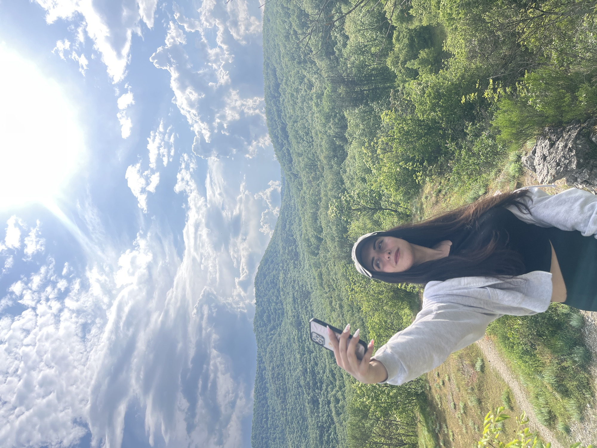 A young woman with long dark hair and fair skin is taking a selfie with her smartphone. She is wearing a gray hooded sweatshirt and dark top. The background is a lush green mountain covered in trees with a partly cloudy sky. The location appears to be an elevated area near Kamchia, Bulgaria, given the mountainous terrain and abundant vegetation consistent with the region's geography.
