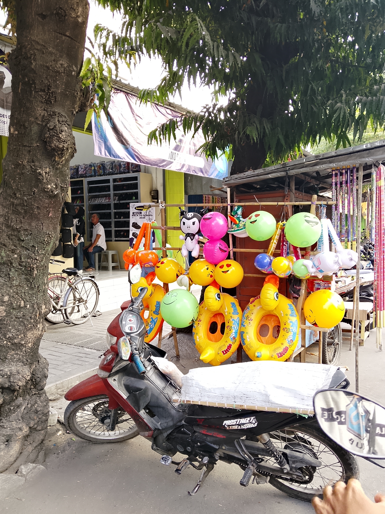 A red and black motorcycle is parked on a paved street in front of a store selling inflatables and other items. In the background, a man is sitting on a stool inside the store. A bicycle is parked next to a tree on the left side of the image. The store has shelves filled with shoes and bags. Various inflatable toys, including smiley faces and animal-shaped rings, are displayed outside the store. The scene appears to be a street market or sidewalk vendor in Balun, Indonesia.
