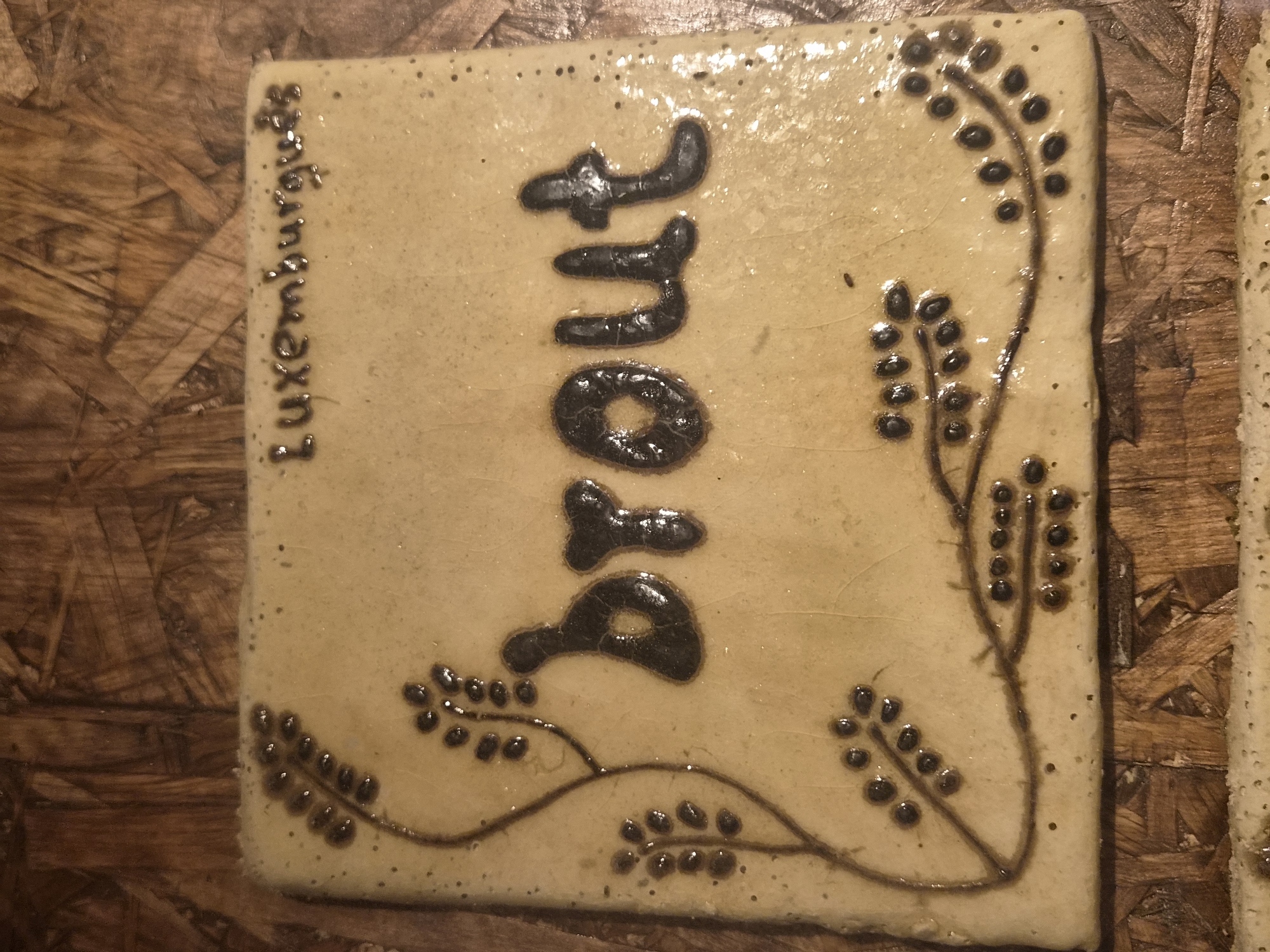The image shows a rectangular ceramic tile with a light beige, slightly crackled glaze, resting on a wooden surface. The tile has decorative elements and text. On the upper right corner of the tile, the word "Luxemburguds" is inscribed in dark brown lettering. The center of the tile features the word "brout" in large, bold, dark brown, raised lettering with a glossy finish. To the left and bottom of the tile, there are thin, vine-like dark brown lines with small, berry-like dark brown dots, creating a botanical motif. The wooden surface appears to be made of pressed wood or particle board with a visible grain pattern. No people or specific location in Seia, Portugal are visible in the image.
