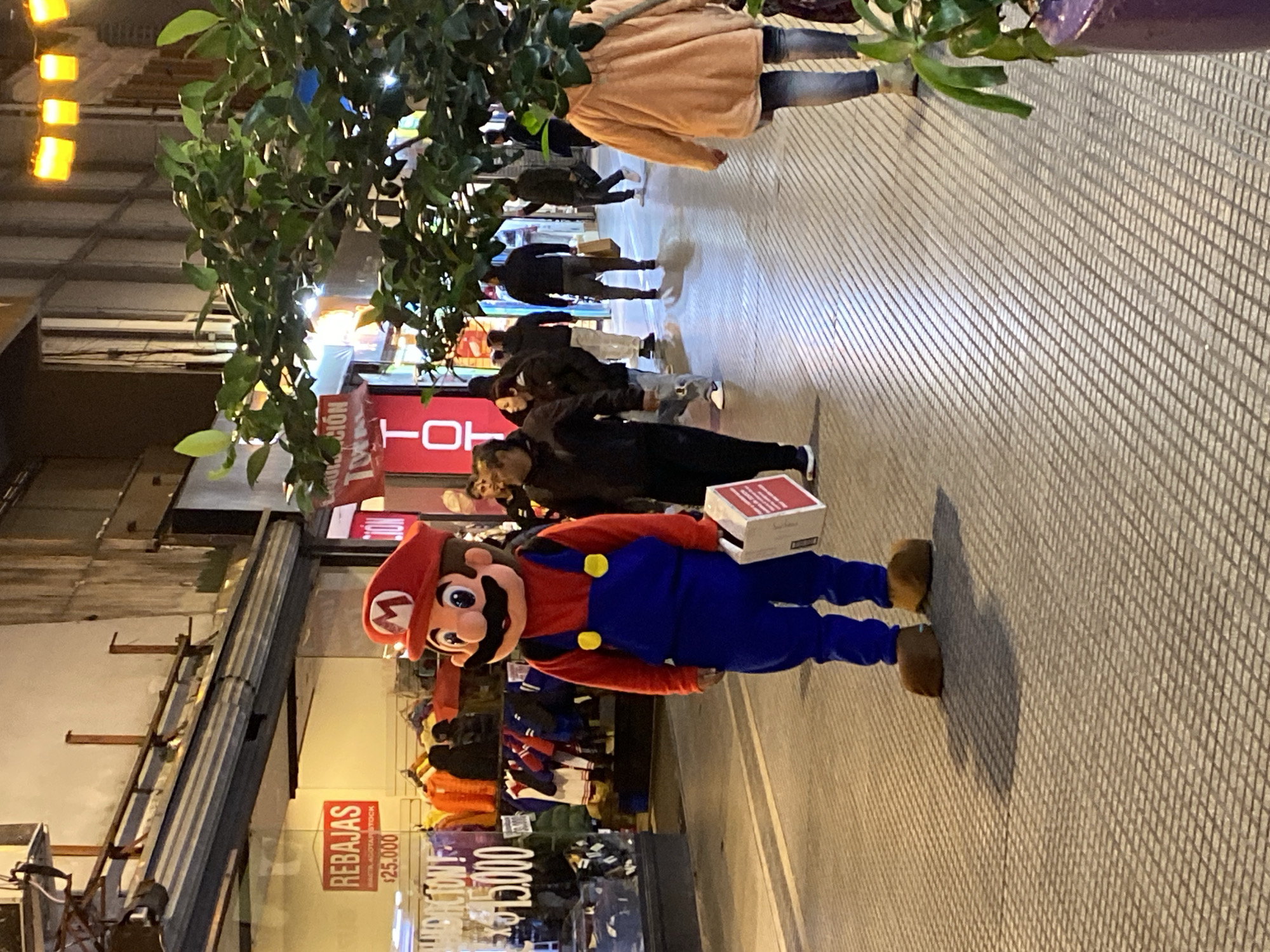 A full body shot of a person in a Mario mascot costume, standing on a sidewalk in Buenos Aires, Argentina. They are holding a white box. Other pedestrians are walking in the background. There is a tree to the left and storefronts with clothing on display. A “REBAJAS” sign is visible on a storefront. The sidewalk is patterned with a grid-like texture. Street lighting is visible in the background.