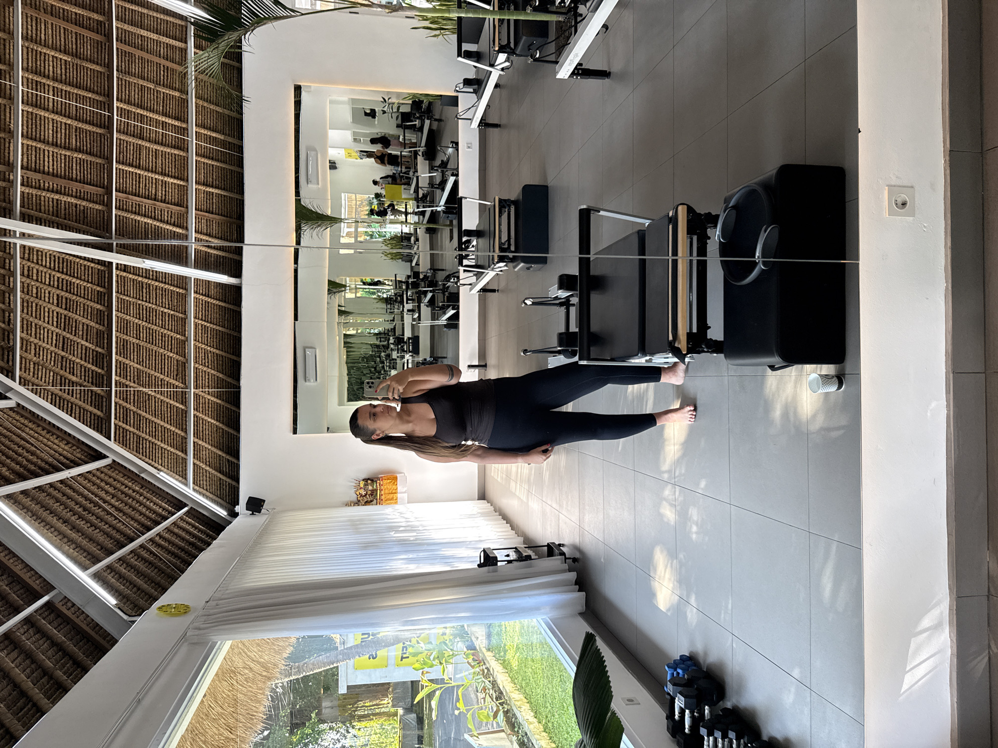 The image depicts the interior of a fitness studio, likely in Ubud District, Indonesia, as suggested by the architectural style. The interior space features a thatched roof, suggesting a tropical location. The floor is tiled in a light color, and natural light streams in through a large window. Exercise equipment, including Pilates reformers, is visible in the foreground and in a mirror on the wall. A woman in athletic wear is positioned in the center of the frame, presumably taking a selfie, holding a smartphone. Other indistinct individuals can be seen reflected in the mirror, possibly exercising. Dumbbells are visible near the window. The color palette is predominantly neutral, with white walls and gray flooring.