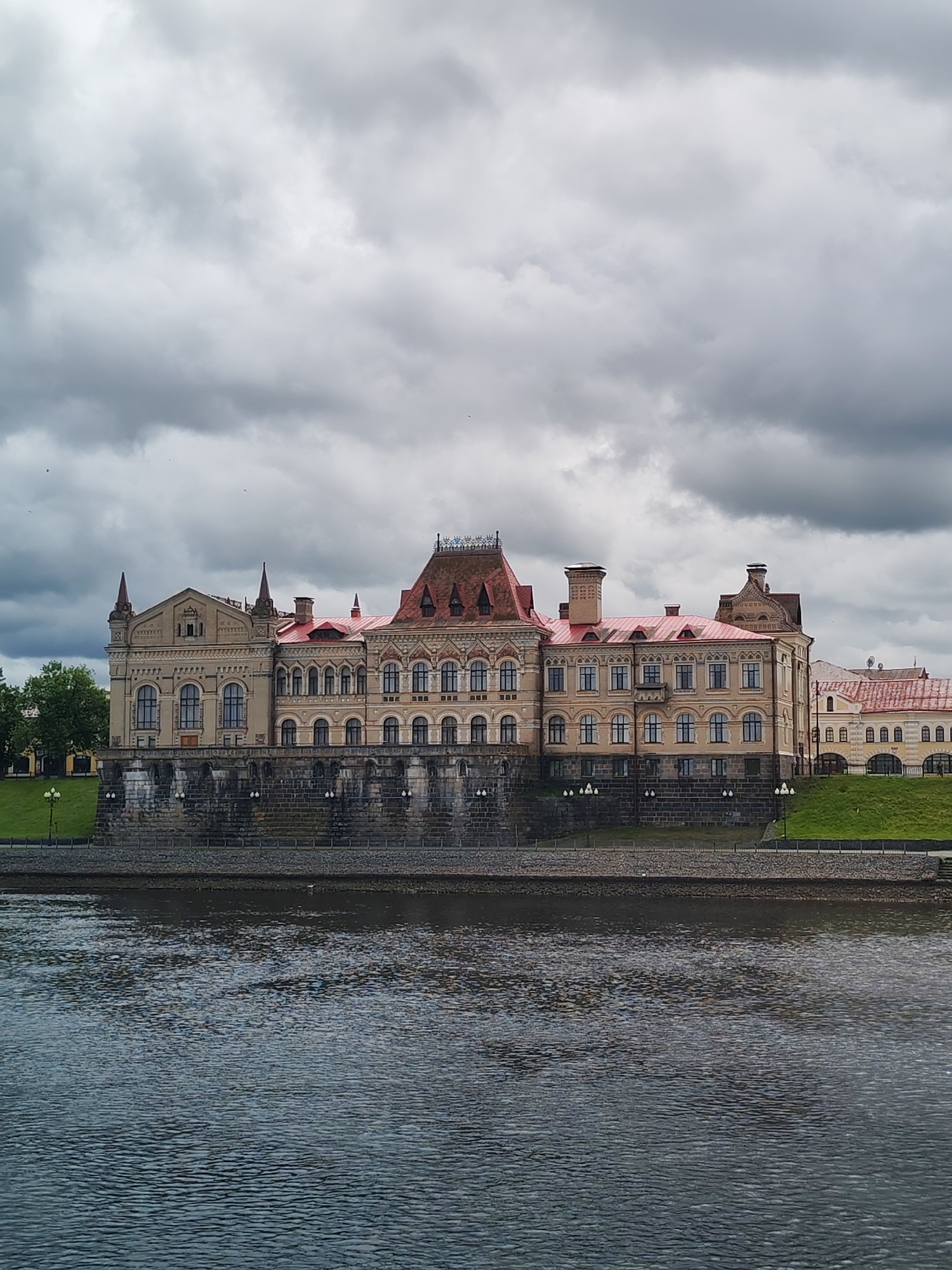 The image depicts the Rybinsk State History, Architecture and Art Museum in Rybinsk, Russia, as seen from across the Volga River. The museum, a large beige building with a red roof, rests on a stone embankment. A row of lampposts lines the embankment in front of the museum, and a small patch of green grass is visible to the right. The river occupies the lower portion of the image, showing the water's texture. Overcast skies fill the upper half of the image, suggesting a cloudy day. There are no people visible in the shot.