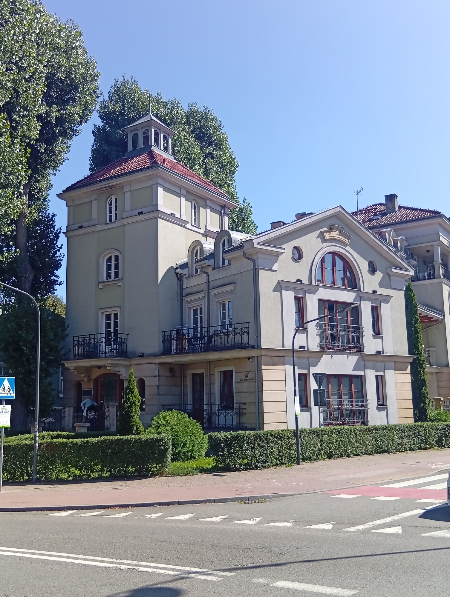 The image depicts a sunny day in the city of Sopot, Poland, showing a pale green, multi-story building with a red-tiled roof and a small tower with a bell-shaped cupola. The building features multiple windows, some with arched tops, and decorative balconies with wrought-iron railings. Adjacent to the main building, another similar structure is partially visible. The building is situated behind a neatly trimmed green hedge, which separates it from a sidewalk and a street. The street has white triangular markings and a red-and-white pedestrian crossing. A street lamp and a blue and white traffic sign indicating pedestrian and bicycle crossing are visible near the building's entrance. A person in a light-colored shirt and dark pants is standing near the entrance of the building. Tall, leafy trees frame the left side of the building. The sky is clear and blue.