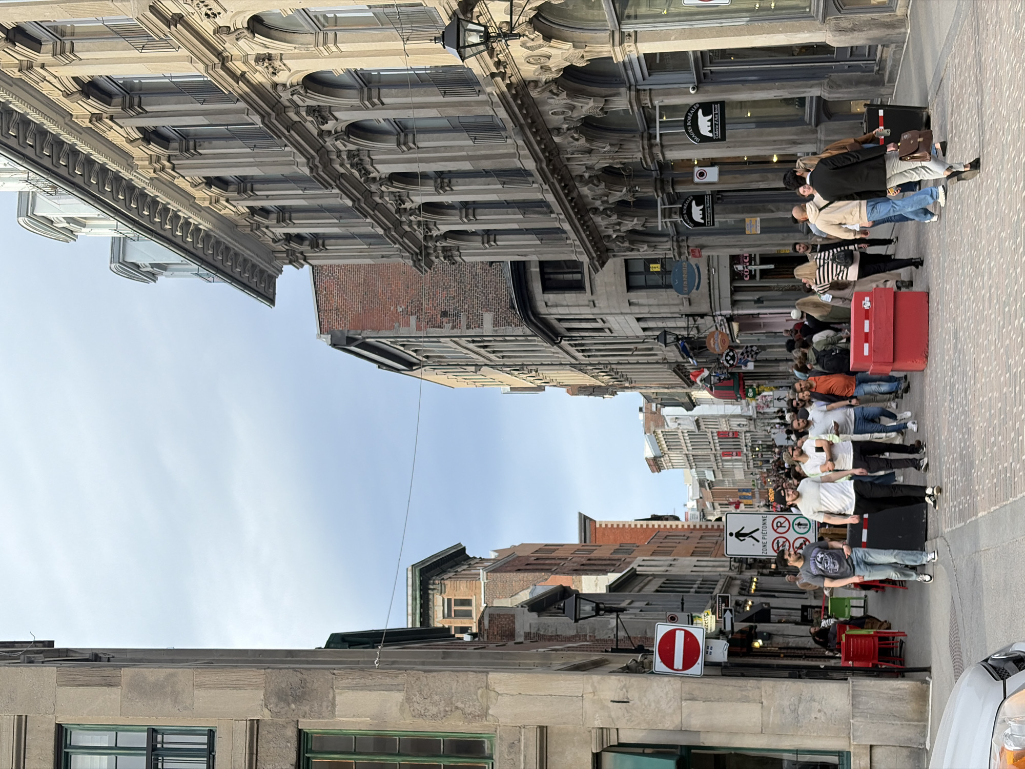 An eye-level, outdoor photograph shows an urban street scene in Montreal, Canada. The image has been rotated 90 degrees counter-clockwise. Pedestrians walk along a pedestrian-only street between buildings with decorative architectural details. Several people are wearing face masks. A red mailbox sits along the street. A "No Entry" sign is visible. Pedestrian crossing signs can be seen. A parked car is visible on the right. The sky is light blue with a few clouds.
