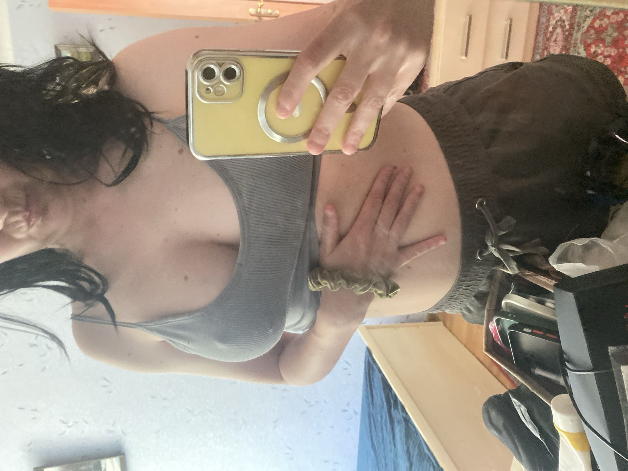 A woman is taking a mirror selfie in a room. She has dark hair and is wearing a gray tank top and dark shorts. She is holding her stomach with her left hand and a yellow iPhone in her right hand. The room appears to be a bedroom, with a bed, dresser, and rug visible. The lighting is natural, coming from a window.