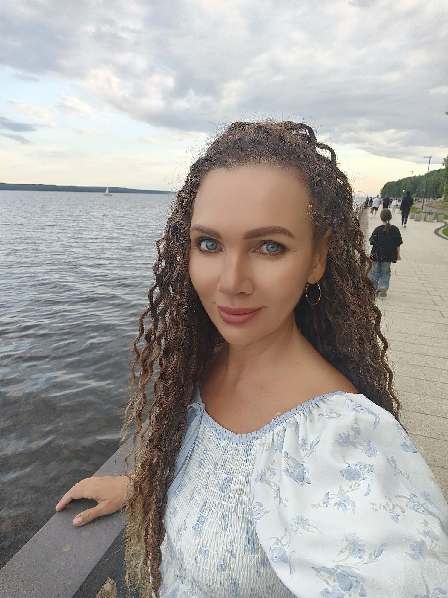 A woman with long, curly brown hair and blue eyes takes a selfie outdoors. She is wearing a white off-the-shoulder top with a blue floral pattern. Her left hand rests on a dark railing. Behind her, a body of water, a walkway, and a line of trees are visible. Several people are walking on the walkway in the background, and a sailboat is in the distance on the water. The sky is overcast with gray clouds. The location appears to be the embankment in Chaikovsky, Russia, possibly near the Kama River.