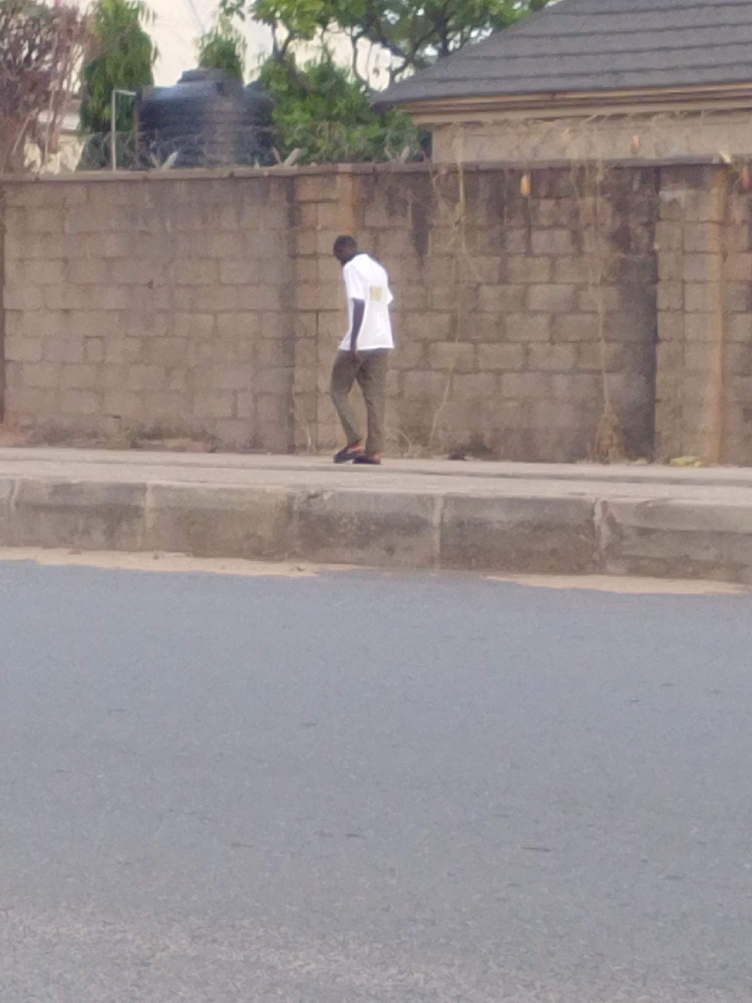 The image shows a man walking on a sidewalk in Bauchi, Nigeria. He is wearing a white shirt and olive-green trousers. He is walking along a grey brick wall topped with barbed wire. Behind the wall are trees and a water tank. The sidewalk is concrete, and the road is asphalt. The scene appears to be a residential area in Bauchi.