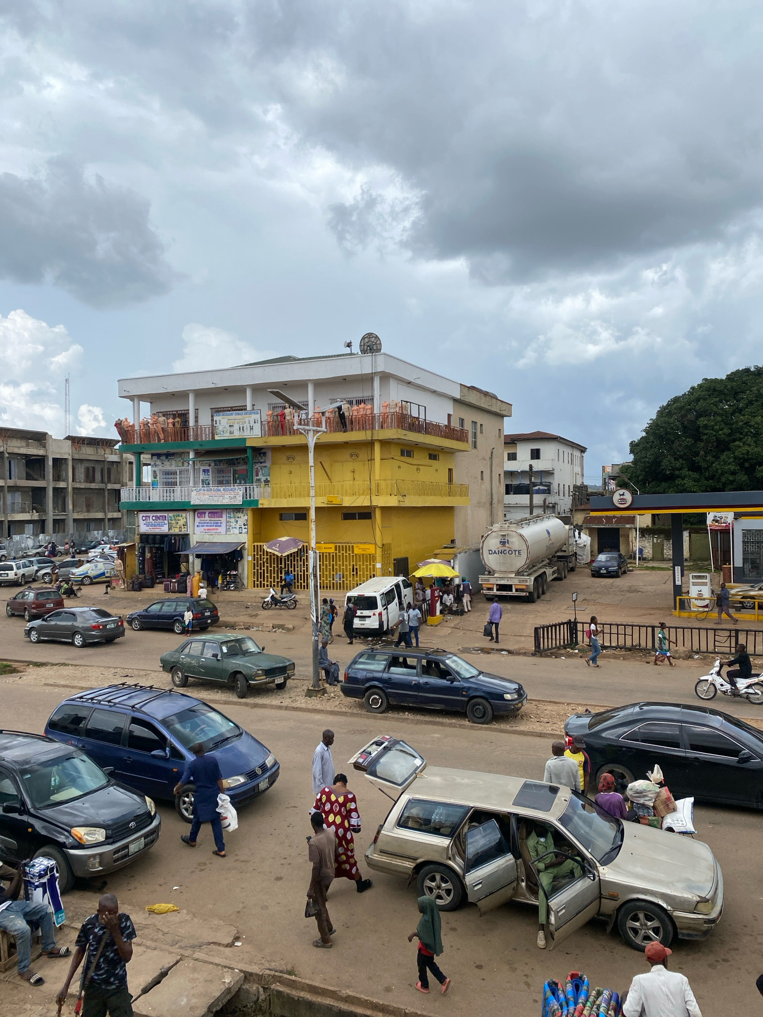 This image depicts a busy street scene in Jos, Nigeria, characterized by a mix of commercial buildings, vehicles, and pedestrians. The buildings are a mix of multi-story structures, some appearing new and others under construction, with colorful awnings and signs. One prominent building is painted yellow and features multiple levels with balconies.
Vehicles are numerous and varied, including cars, SUVs, a tanker truck labeled "DANGOTE," and a motorcycle. Several cars are parked along the roadside, while others are in motion. One car has its trunk open, revealing stacked items. A group of people is gathered near a fuel station with a visible "TOTAL" logo.
Pedestrians are seen walking on the sidewalks and crossing the street, some carrying bags. Their clothing is diverse, reflecting a casual and everyday urban environment. The sky is overcast with heavy clouds, suggesting a potentially rainy or humid day. The ground is a paved road with visible cracks and dirt. The overall impression is one of a bustling urban marketplace or transportation hub in Jos.