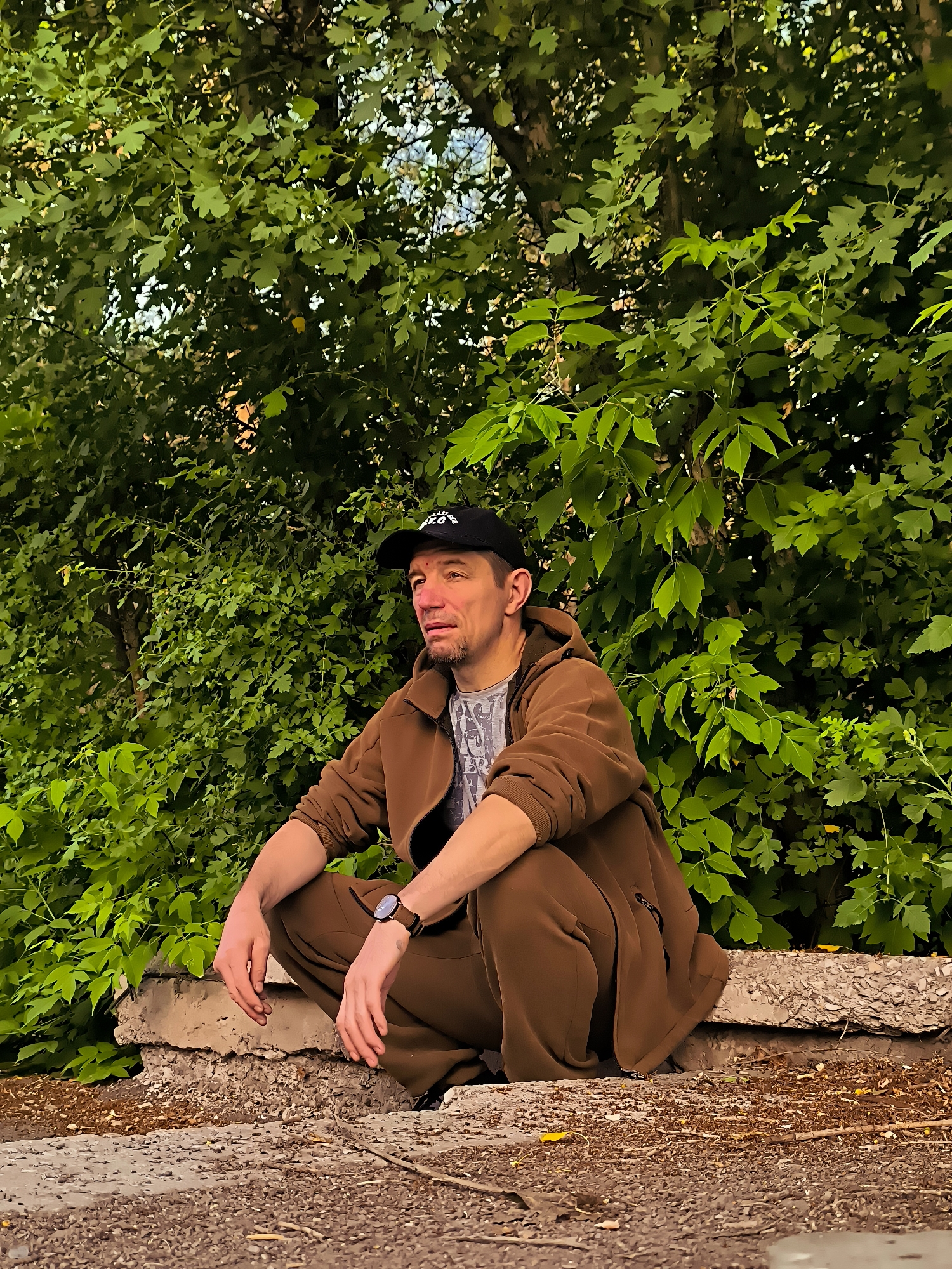 A man wearing a brown tracksuit and a black baseball cap squats on a concrete ledge with lush green foliage behind him. He is looking to his left with a pensive expression. His left arm rests on his knee, and his right arm is bent with his hand near his face. He is wearing a wristwatch on his left wrist. The ground in front of him is covered in dirt and small stones. The scene appears to be outdoors, possibly in a park or a natural area within the city of Qarağandı, Kazakhstan.