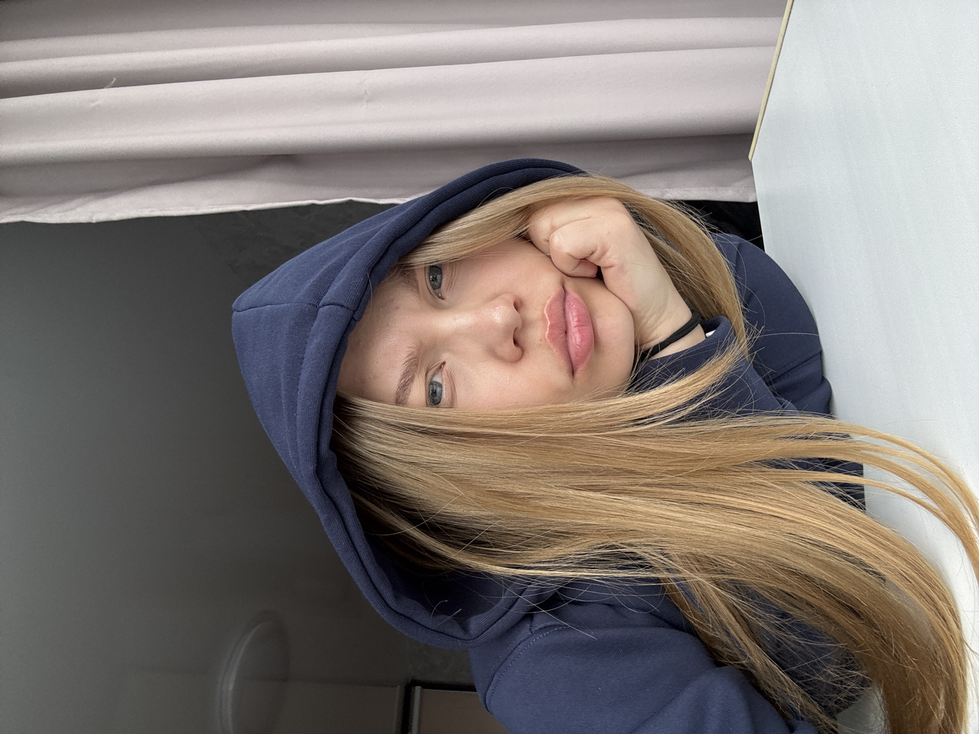 A young woman with blonde hair wearing a navy blue hooded sweatshirt rests her face on her hand. She has light skin, blue eyes, and pink lips. The background includes a light grey curtain, a dark grey wall, and a white wall. The location is the city of Chaikovsky, Russia.