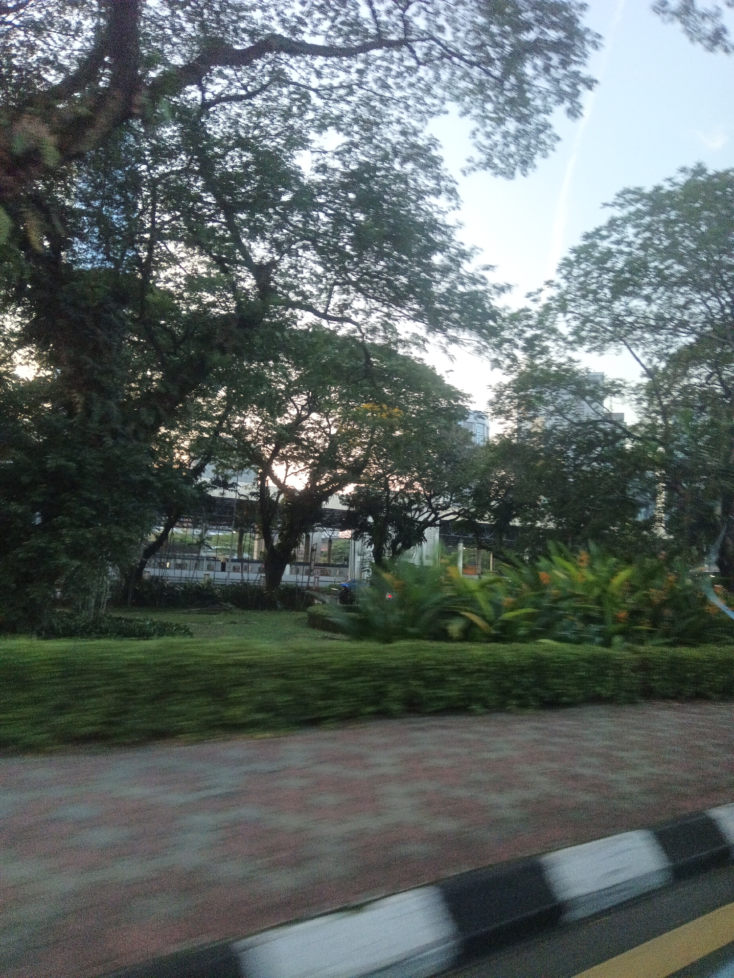 The image captures a motion-blurred view of a park-like area with lush green grass, dense trees, and tropical foliage. In the background, a modern building with glass windows is visible, partially obscured by the trees. The foreground shows a brick pathway and a black and white striped curb, indicating a road or sidewalk. The lighting suggests it is either early morning or late afternoon, with soft sunlight filtering through the trees. The scene is devoid of people. This location appears to be in Kuala Lumpur, Malaysia, possibly near a green space or park adjacent to an urban area.