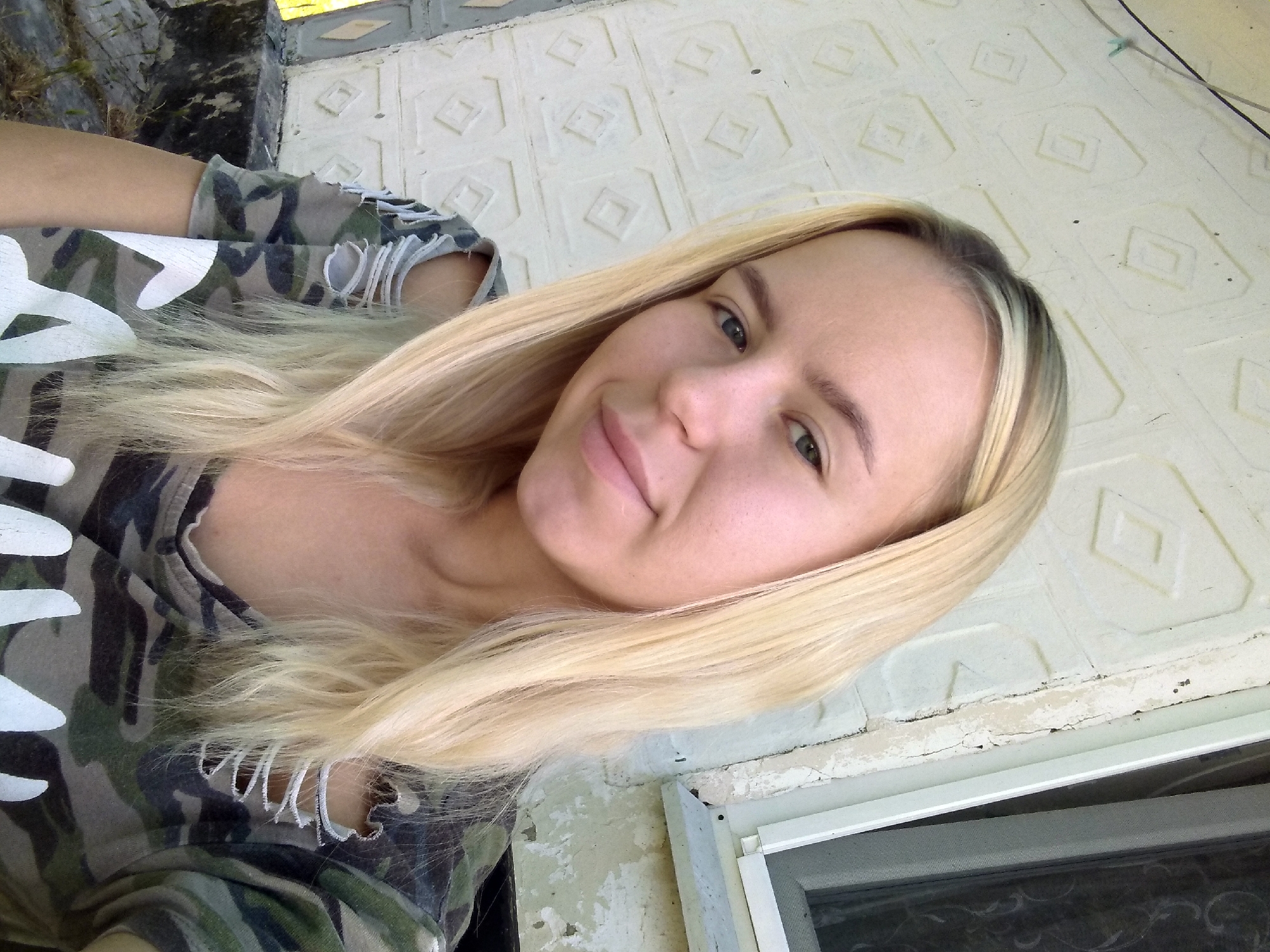 A young blonde woman with light skin and blue eyes is taking a selfie outdoors. She is wearing a camouflage t-shirt with distressed shoulder cutouts and a large white logo on the front. The background consists of a textured, off-white wall with a diamond pattern and a white-framed window on the right side. The lighting suggests it is daytime. The location is the city of Radisne, Ukraine.