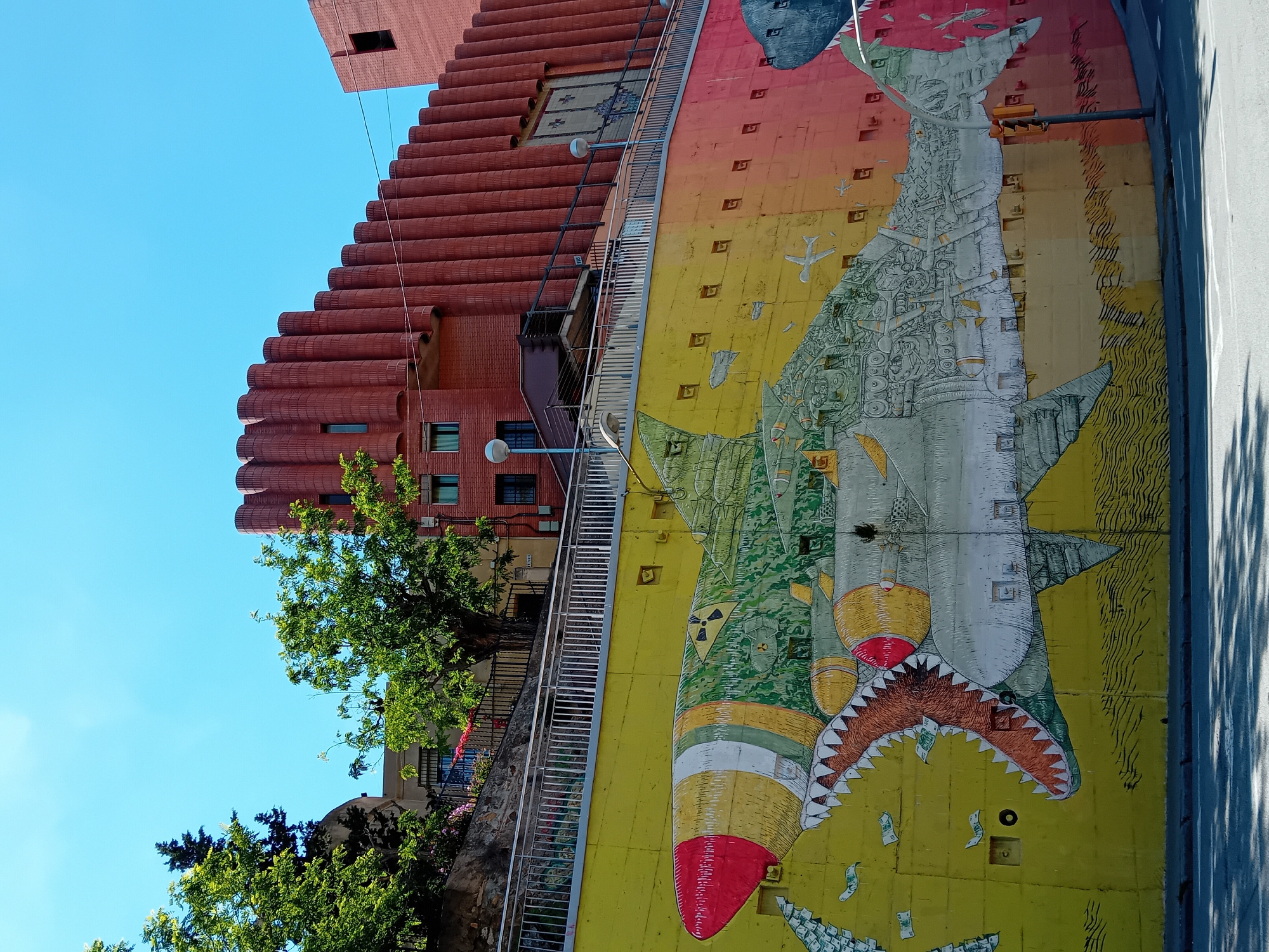 The image features a colorful mural painted on a wall, likely in Barcelona, Spain, given the architectural style of the building next to it. The mural depicts a large shark made of missiles and money, with a background of bombs and dollar bills. The shark's head is a large missile with a red nose cone and sharp, white teeth. The shark's body and fins are made of missile parts and mechanical drawings. The mural is painted on a yellow background that fades into orange near the top.
To the left of the mural is a tall, red-brick building with a unique, terraced design. A staircase runs along the wall between the mural and the building. Some trees are present in front of the building. The sky is blue and clear above the building. The photo seems to be taken during a sunny day.