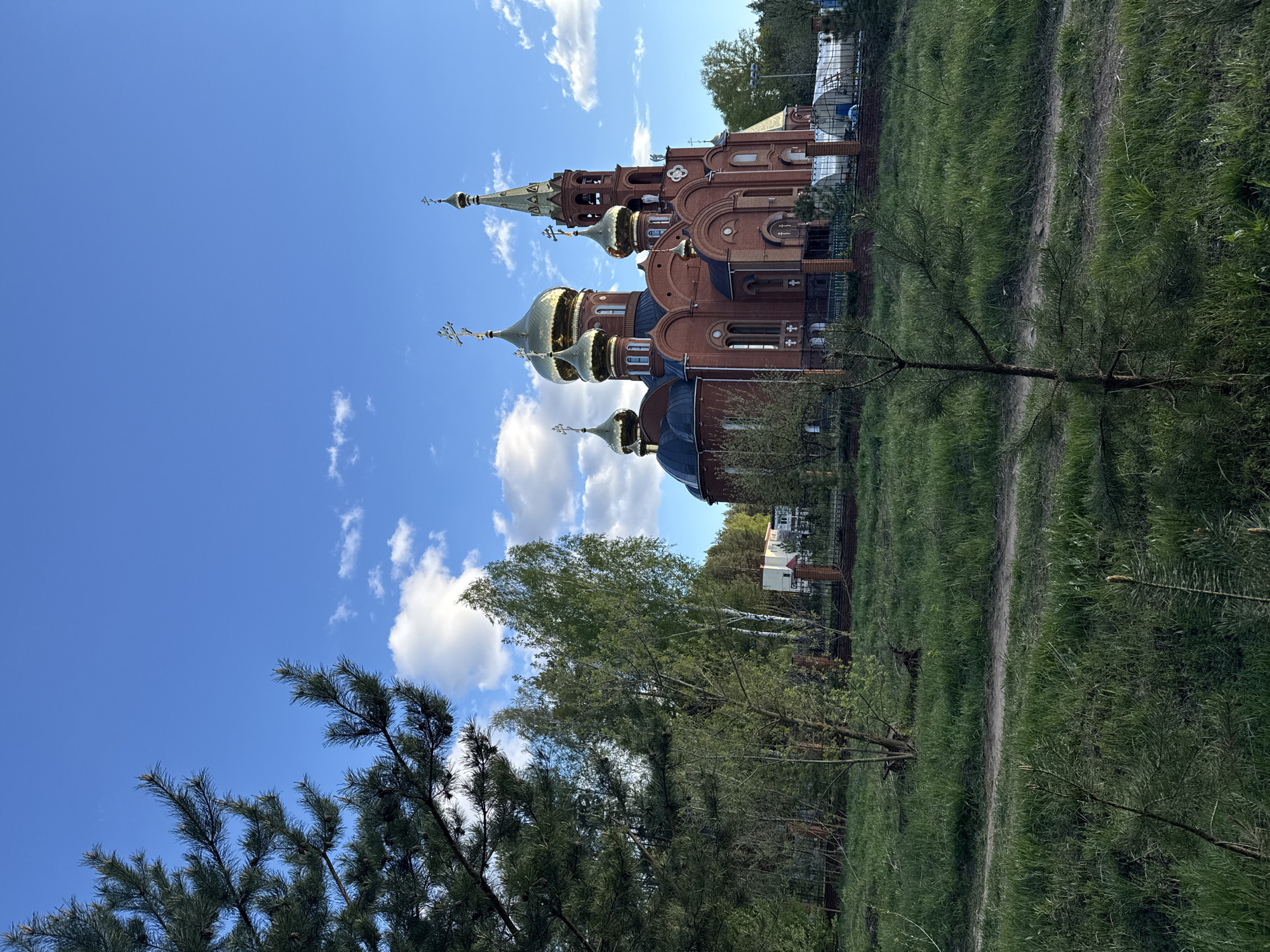 The image shows an Orthodox church in an unknown city. The church is brick-red, with golden domes. It is situated on a green, grassy area with trees. The sky is blue with some white clouds. A narrow dirt path leads to the church. A pine tree is prominent in the foreground.