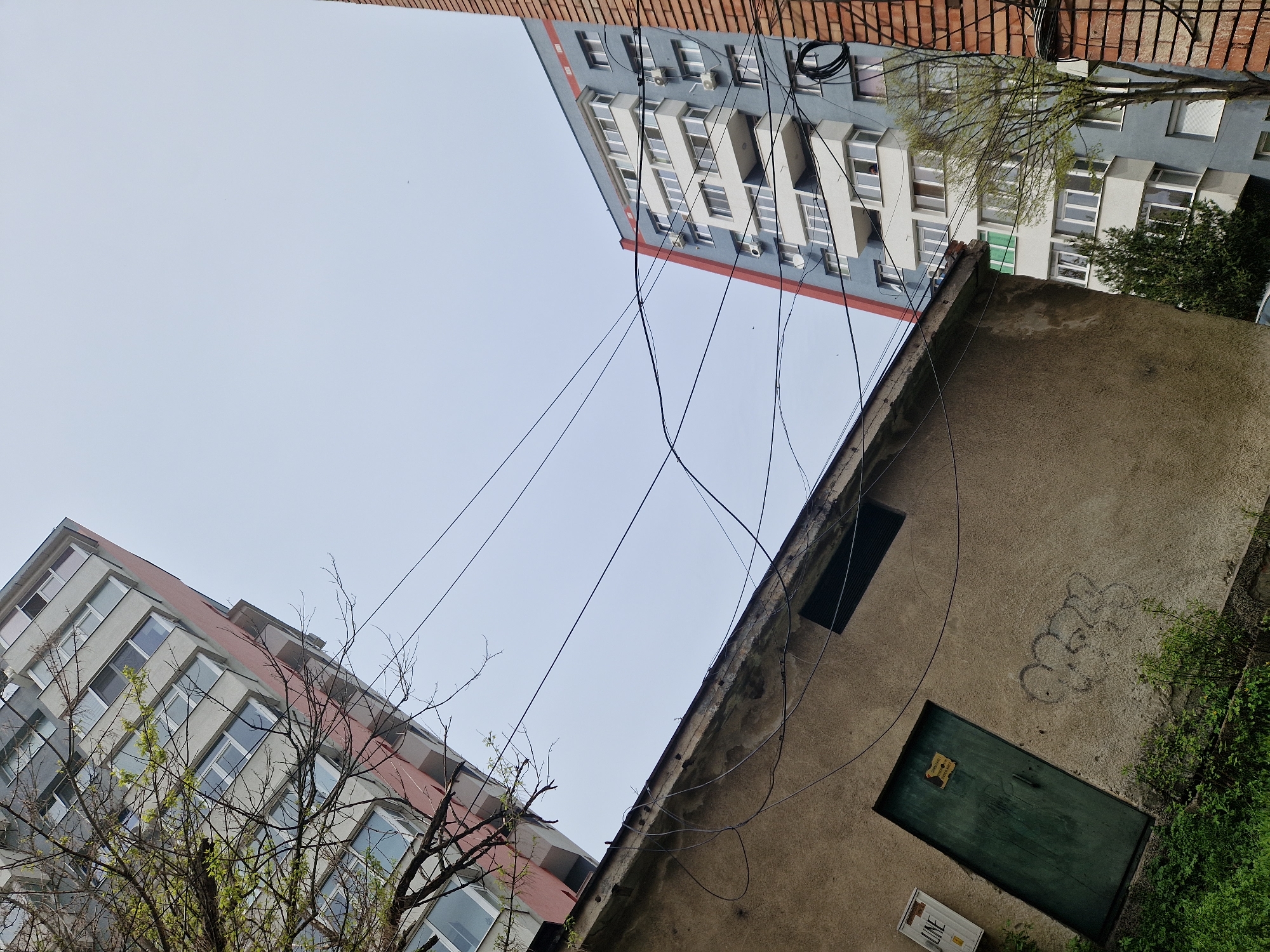 The image depicts an urban scene in Lovech, Bulgaria. It shows a low-angle view of two buildings, one painted grey and white and the other with a red brick top. A smaller, weathered building with a green door is in the foreground. Power lines stretch across the sky between the buildings. There is a patch of graffiti on the wall of the smaller building. Greenery is visible around the buildings, particularly near the green door. The sky is overcast.