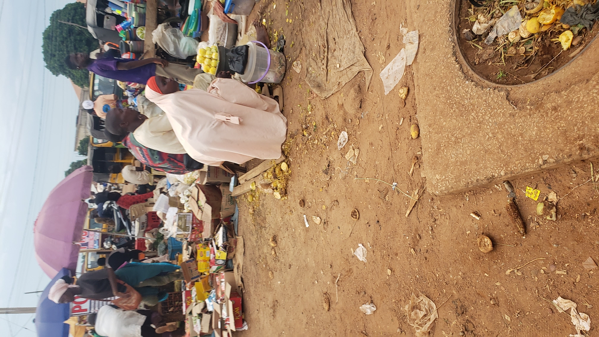 The image depicts a bustling market scene in Kakuri Gwari, Nigeria. The ground is predominantly dirt, scattered with debris, including discarded food items like corn husks and fruit peels, as well as plastic wrappers and bags. In the foreground, a concrete structure with an open manhole filled with organic waste is visible. Several individuals are present, engaged in market activities. A person wearing a light pink hijab is crouched down near a pile of yellow fruits, possibly lemons, and a black plastic bucket. Other people are seen standing and moving around the market, with some behind stalls laden with various goods. Umbrellas, including a large purple one, provide shade. The background shows more market stalls, vehicles, and trees, indicating a typical urban marketplace environment under an overcast sky.