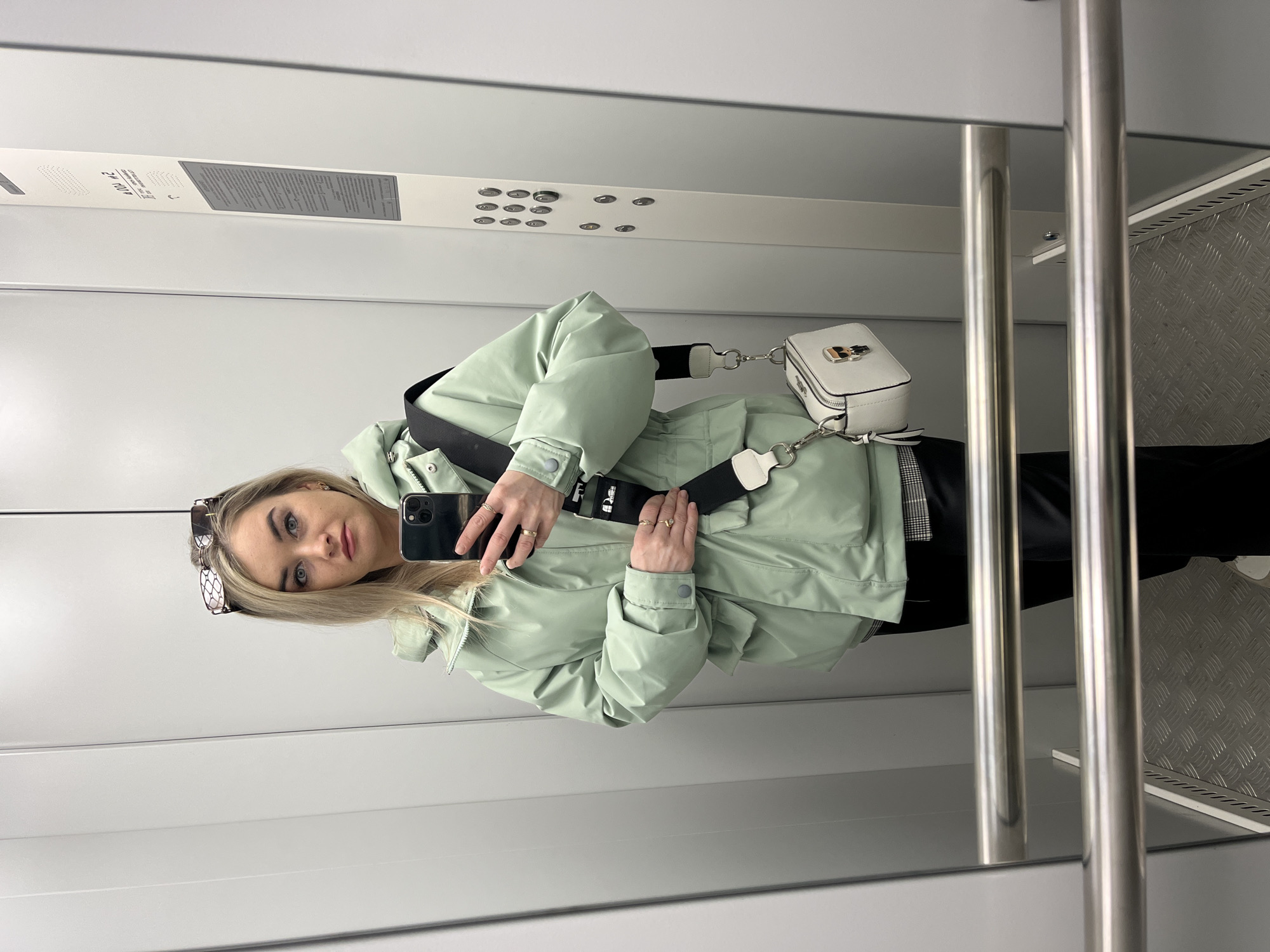 An adult female is depicted taking a mirror selfie inside an elevator in Chaikovsky, Russia. She has blonde hair, blue eyes, and fair skin, with sunglasses resting on her head. She is wearing a mint green hooded puffer jacket with a drawstring waist, dark pants, and a plaid undershirt. Gold rings adorn several fingers of her right hand, which is holding the jacket, while her left hand holds a dark-colored smartphone (consistent with an iPhone model with three rear cameras) towards the mirror to capture the image. A white rectangular crossbody bag with a black and white striped strap, featuring a "Karl Lagerfeld" emblem, hangs from her left shoulder. The elevator interior features light grey, reflective wall panels. The floor is made of metallic diamond plate. On the right, a vertical control panel displays multiple round silver buttons and Russian text indicating a "Load capacity: 400 kg / 5 persons," "Manufacturer: JSC Moslift," and an "Emergency communication service." Two horizontal, polished metal handrails are visible in the foreground.