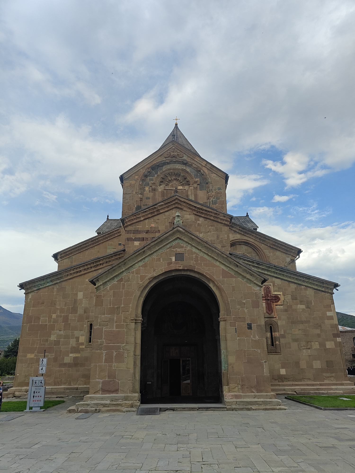 A low-angle, full-front shot captures a grand, ancient-looking stone church or cathedral under a partly cloudy sky. The impressive edifice is constructed from large, brownish-grey hewn stone blocks, giving it a sturdy and historic appearance. Its central feature is a large, deep arched entrance at the base, leading into a dark interior. Above the entrance, the facade rises in stepped gables, culminating in a conical dome topped with a golden cross. Intricate stone carvings adorn various architectural elements, including the arches, window frames, and roof lines. To the right of the main entrance, a prominent red cross motif is carved into the stone wall.
The foreground is a wide, paved area of grey flagstones, leading directly to the church's entrance. Green grass patches flank the paved path. On the left side, near the entrance, a white information sign stands upright. It displays several black pictograms indicating rules or guidelines: a person walking (possibly indicating a path), a wheelchair user, a crossed-out speaker (no loud noise), a crossed-out cigarette (no smoking), a crossed-out dog, a crossed-out flash (no flash photography), and crossed-out food/drink items. Below these symbols, visible Georgian text reads, "გთხოვთ ნუ შედიხართ" (Please do not enter/do these actions), "მიკროფონით" (with a microphone), and "ფლეშ განათებით" (with flash lighting), outlining prohibited activities.
In the far left, beyond the sign, a few blurry figures are visible, likely visitors or parishioners. Distant mountains or hills can be seen on the left horizon. The sky is a mix of light blue patches and soft white clouds. The lighting suggests daytime, possibly late morning or early afternoon, with diffused light that highlights the texture of the stone.