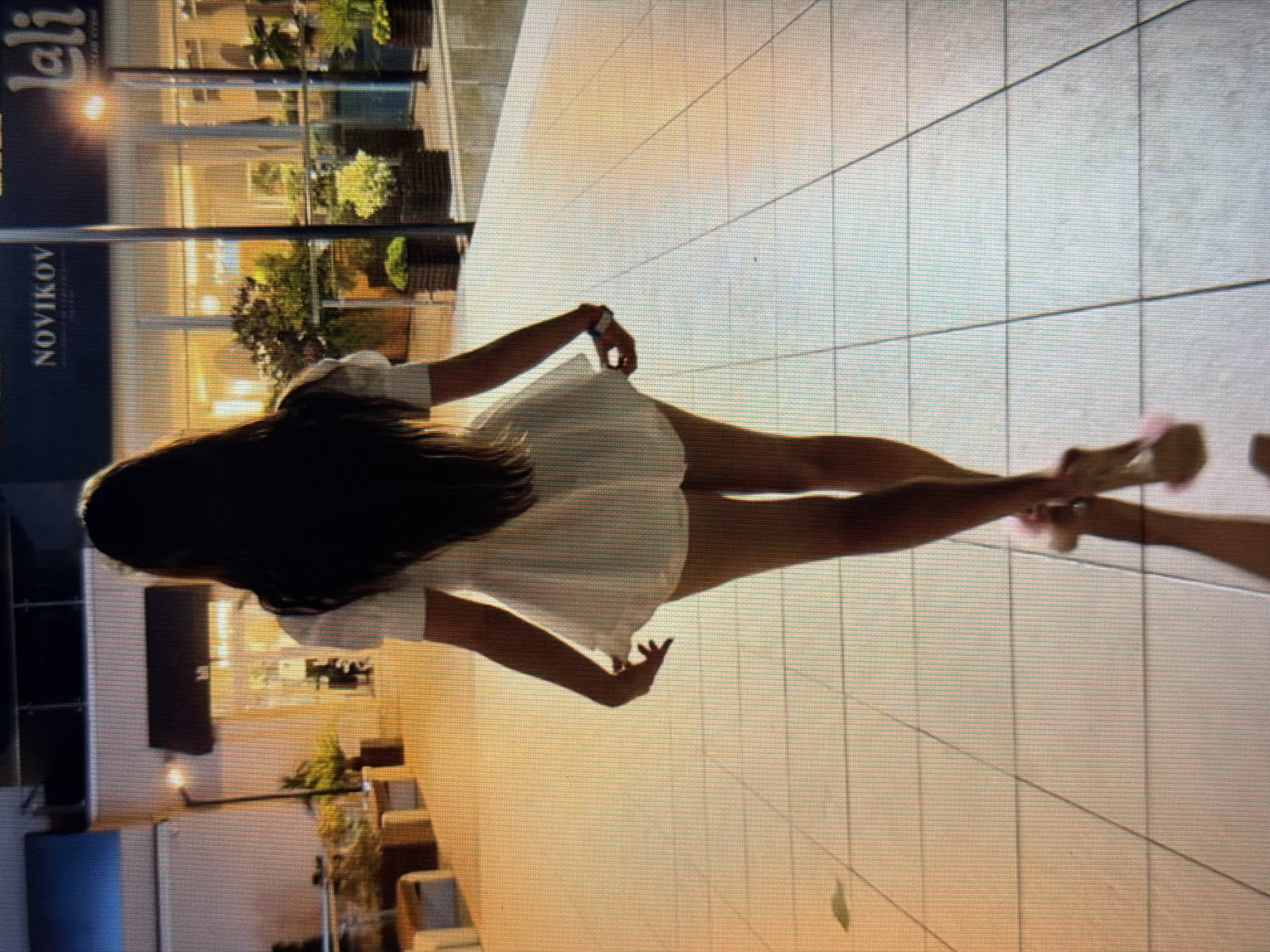 A full-body shot from behind shows a woman walking away from the camera on a tiled walkway at night. The woman has long, dark hair that cascades down her back. She is wearing a short, white, A-line dress with puffed sleeves. Her legs are long and slender, and she is wearing light-colored high-heeled shoes. She is lifting the hem of her dress slightly with her right hand.
The background suggests an outdoor urban environment. To the left, there are some benches and potted plants. To the right, there is a building with large windows and illuminated signs. One sign reads "NOVIKOV" and another appears to say "lali". Streetlights cast a warm glow on the scene, creating reflections on the wet-looking tiled ground. The overall impression is of a chic, upscale location, possibly a shopping area or restaurant district. The location is identified as Sirius, Russia.