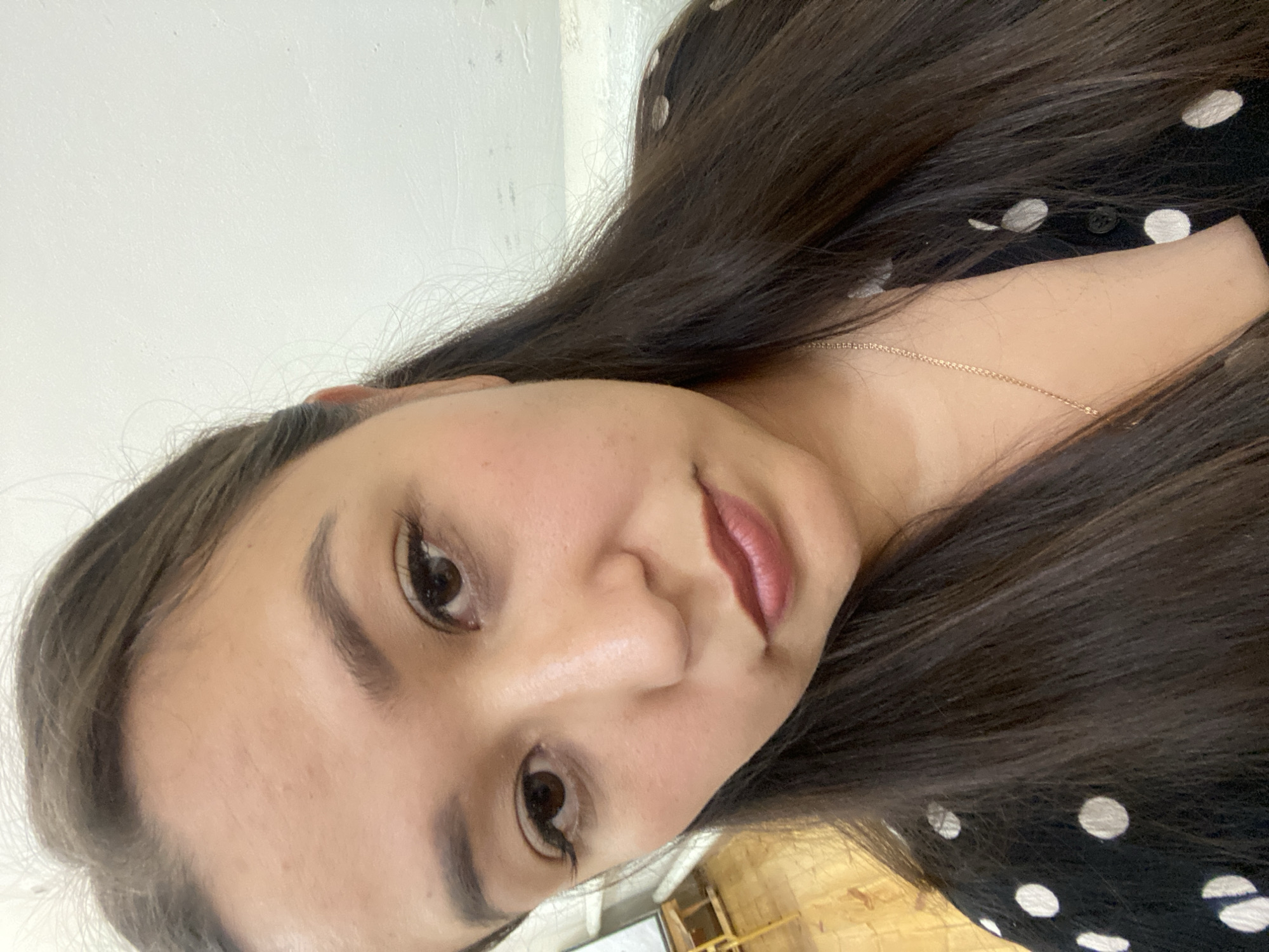 A close-up selfie shows a woman with long, dark hair pulled back from her face, against a plain, light-colored wall. She is wearing a black garment with white polka dots and a delicate gold necklace. Her makeup includes defined eyebrows, eyeliner, mascara, and a muted berry-toned lipstick. Her expression is neutral, with her gaze directed slightly downwards. The background is blurred, but hints of a wooden floor and a light-colored object are visible. The location is Maloarslangulovo, Russia.