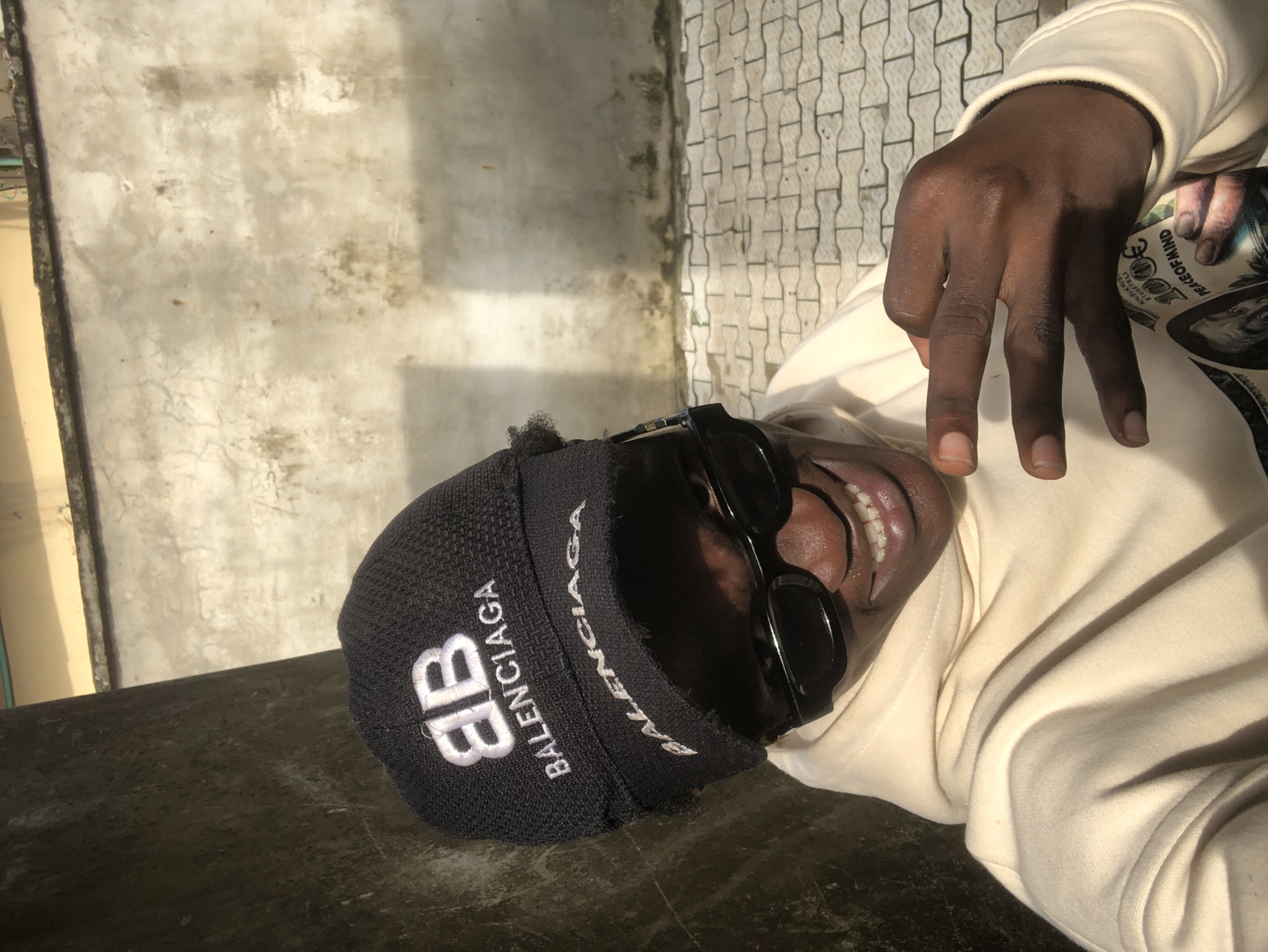 The image presents a close-up, high-angle portrait of an individual, likely male, lying down or leaning back with a broad smile directed upwards towards the camera, revealing white teeth. The person wears a black beanie featuring the white "BALENCIAGA" text and its distinctive "BB" logo prominently on the front. Black sunglasses are pushed up onto their head, and they are dressed in a light cream or off-white hoodie.
In the foreground, one hand is raised, making a gesture with three fingers extended. Partially visible beneath this hand is an object resembling a wallet or small bag, patterned with a dollar bill design. Text on this item includes "PEACEOFMINd" and a stylized "300," likely an altered "100."
The background indicates an outdoor or semi-outdoor setting in Ado, Nigeria. To the left, a textured, weathered light-colored concrete wall is visible. To the right, grey paving stones with a distinct interlocking pattern form another part of the ground or a nearby surface. The lighting is bright, suggesting daylight and sunny conditions, casting subtle shadows. The person appears relaxed and joyful, smiling widely.